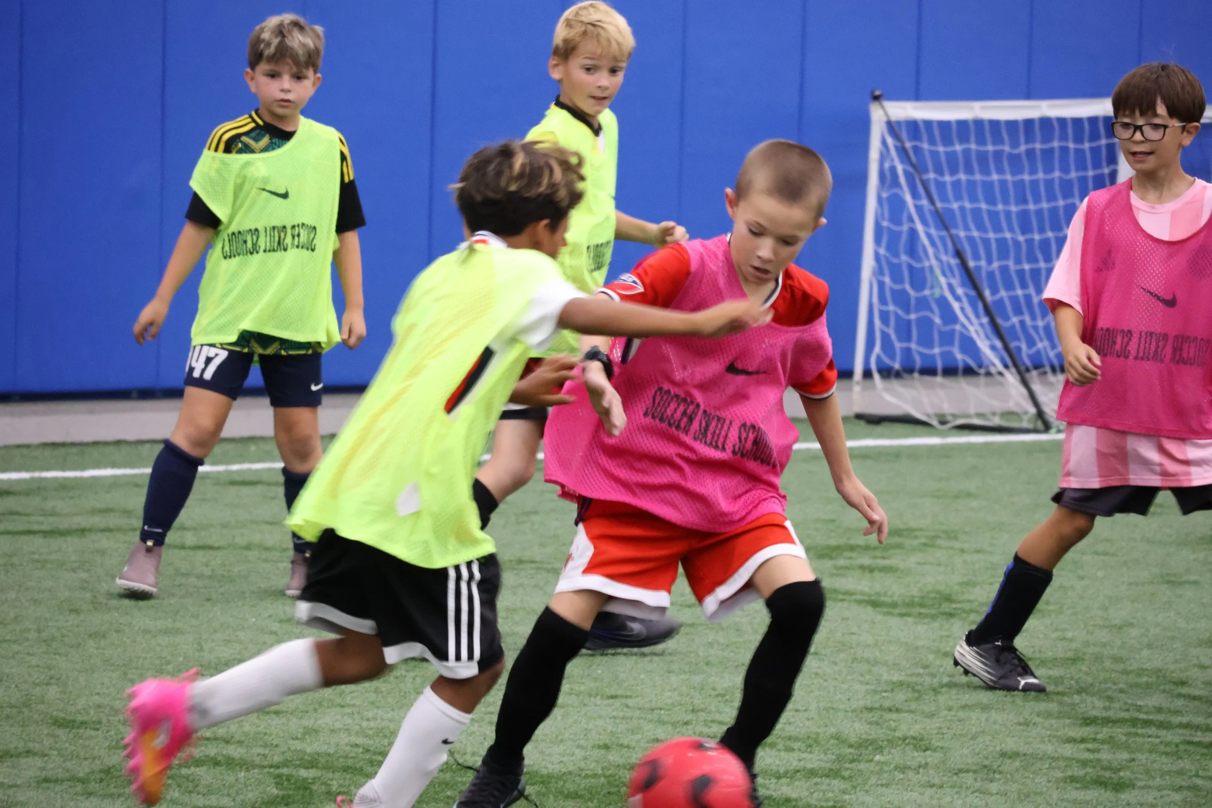 Group of young boys playing soccer indoors on a field with a blue wall and goal net in the background. Some boys are wearing pink and yellow vests, and one boy in a red uniform is kicking a soccer ball.