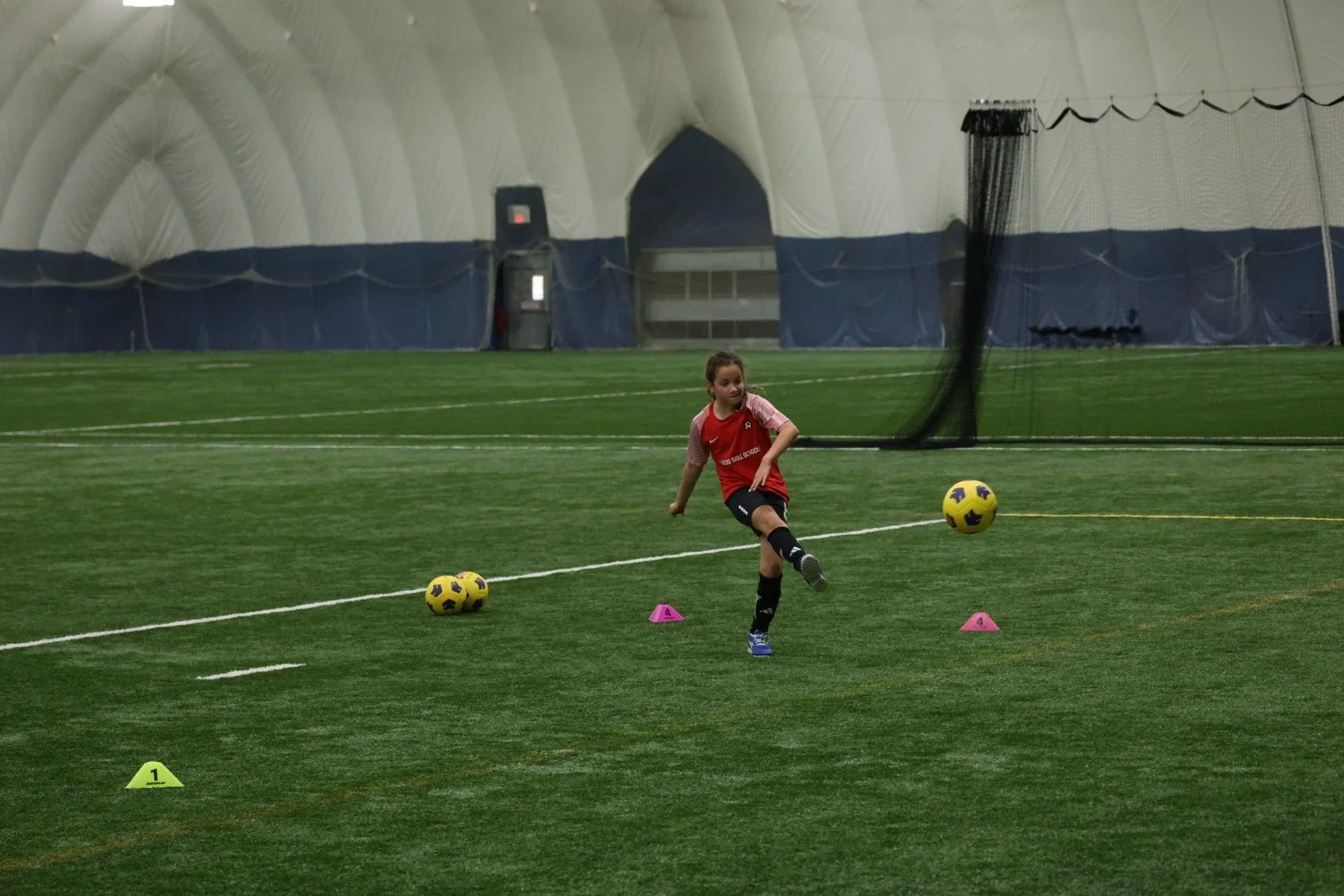 A young girl practicing soccer indoors, kicking a yellow and purple soccer ball. She is wearing a red and pink jersey, black shorts, and black knee-high socks. There are pink and yellow training cones on the green artificial turf field.