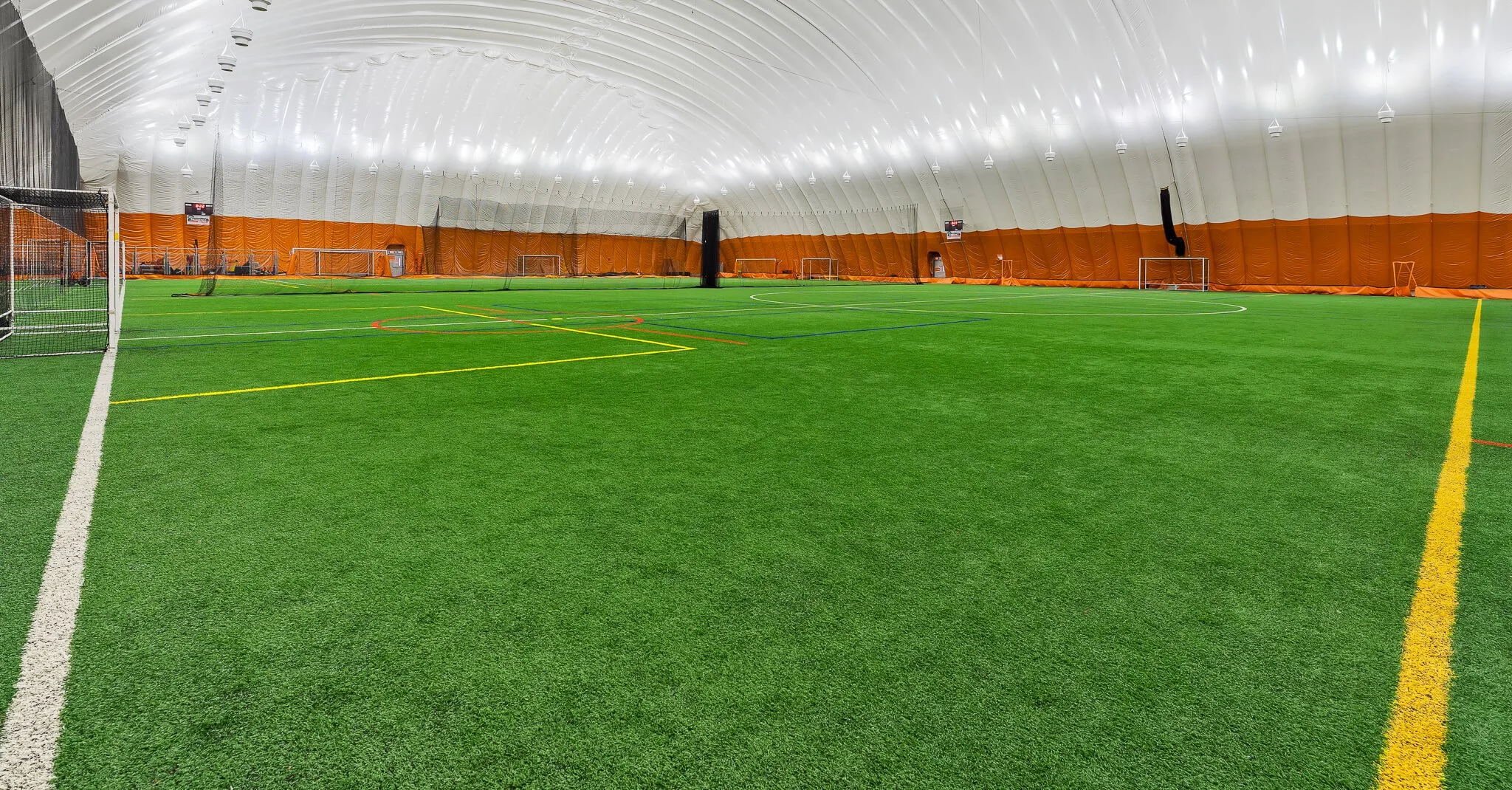 Indoor sports facility with a basketball court and volleyball court, players practicing, and a person walking on the upper walkway. Skill Academy