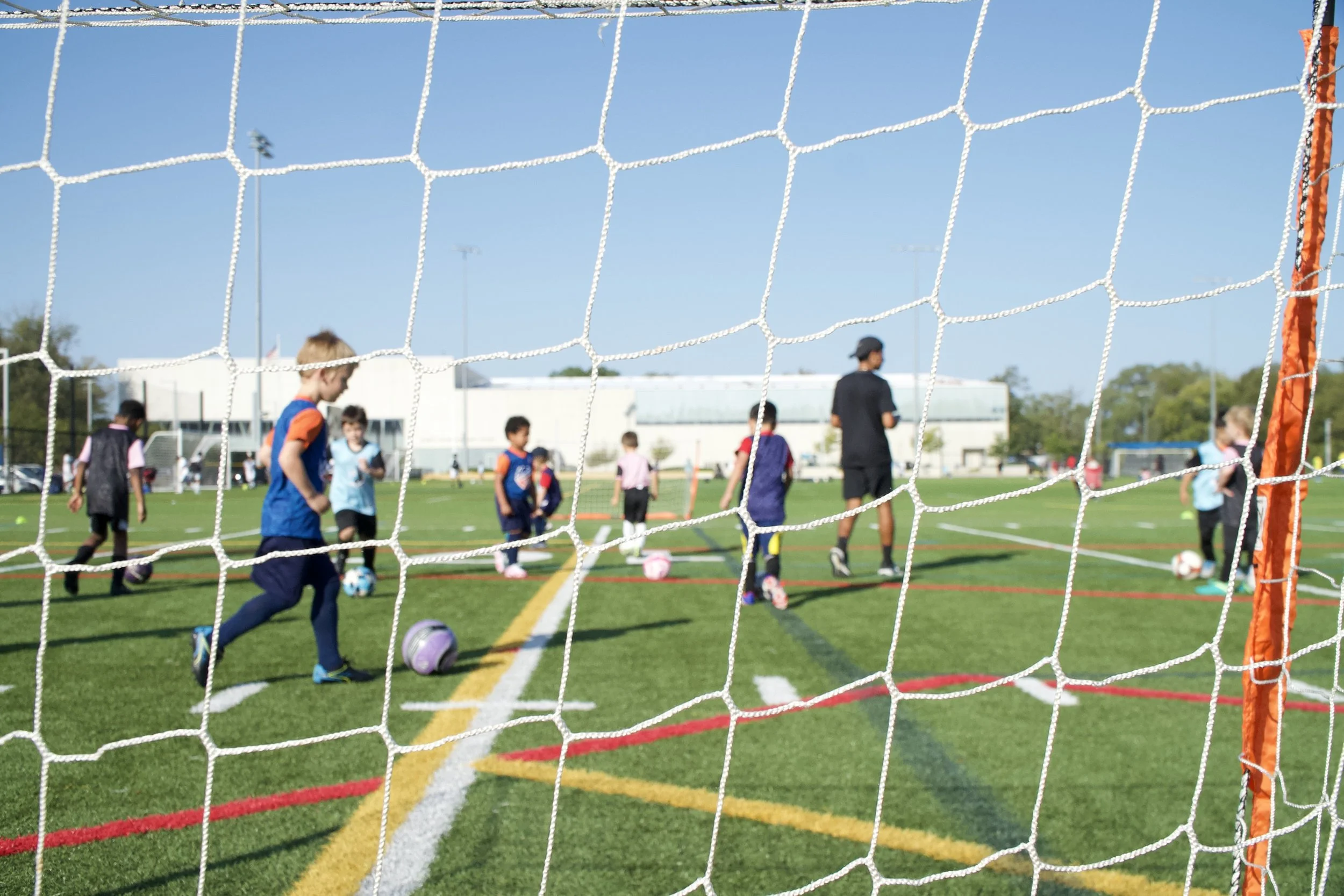 Children in soccer practice on a field, viewed through a goal net, with a coach supervising. The field has colorful boundary lines, and there are buildings and trees in the background under a clear blue sky.