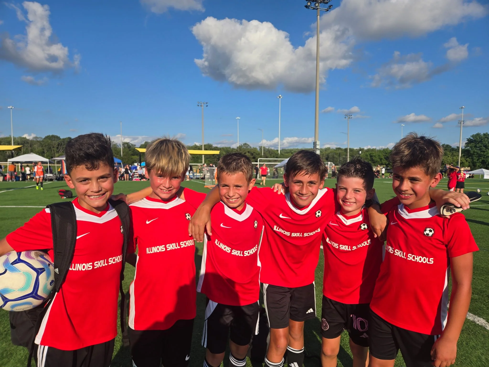 Group of six boys in red soccer uniforms standing on a soccer field with their arms around each other, smiling, in the late afternoon. They are at a soccer tournament, with tents, other players, and spectators in the background under a partly cloudy 