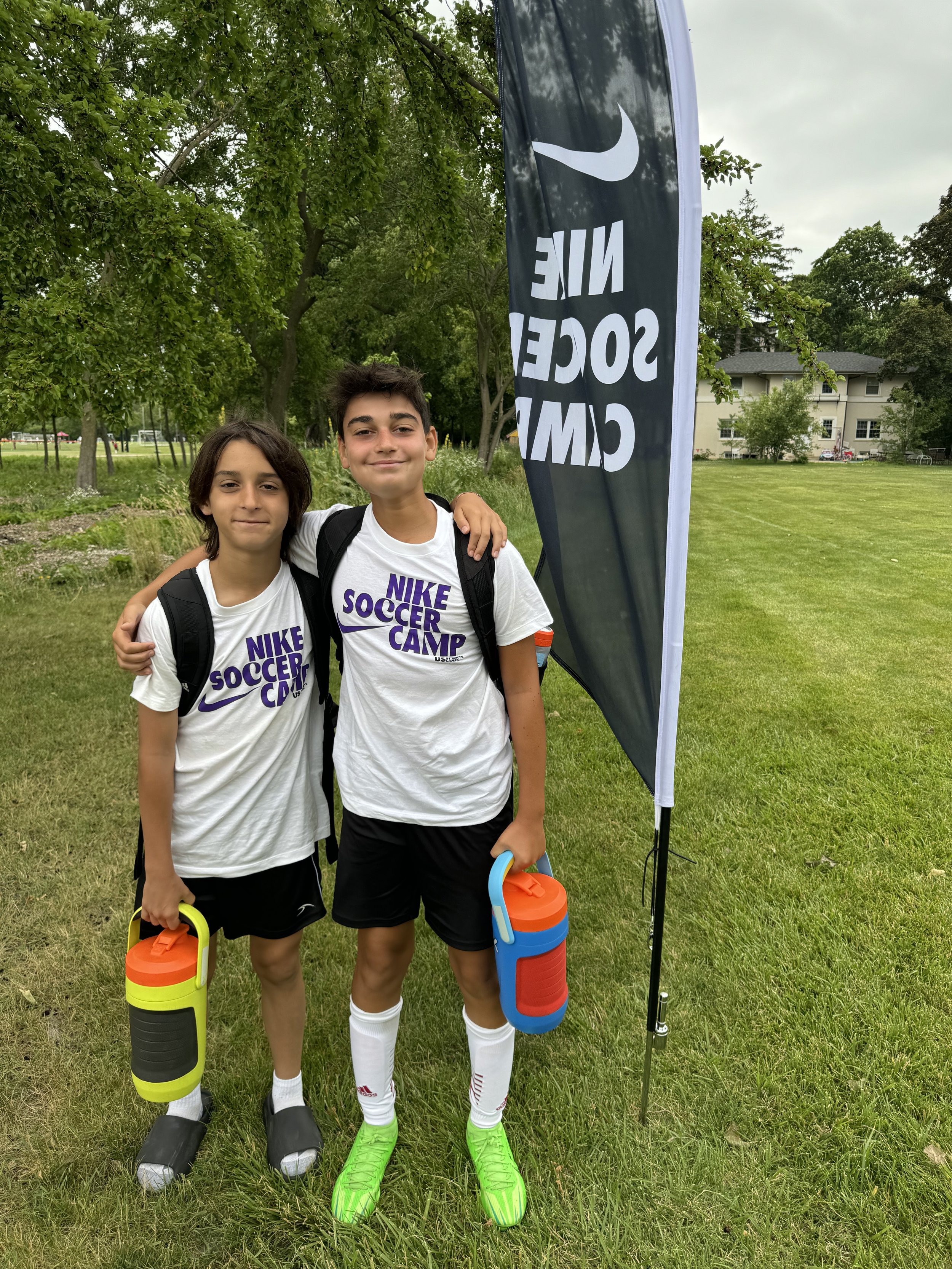 Two boys standing on a grassy field, smiling, wearing white Nike Soccer Camp shirts with black shorts, each carrying a small colorful cooler, with a black sign and trees in the background.