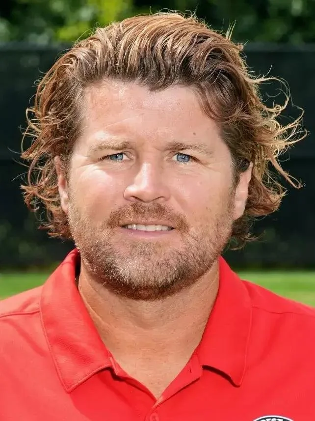 Portrait of a man with curly, reddish-brown hair and blue eyes, wearing a red polo shirt, standing outdoors with greenery in the background.