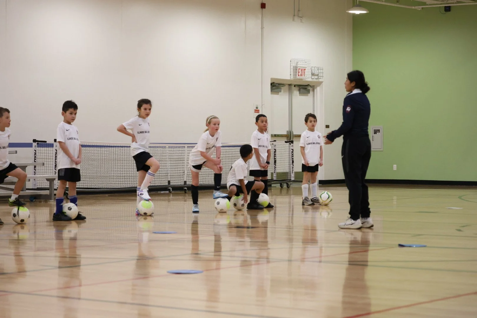 Children in a gymnasium participating in a soccer training session led by an instructor. Children are standing and sitting with soccer balls at their feet, practicing skills.