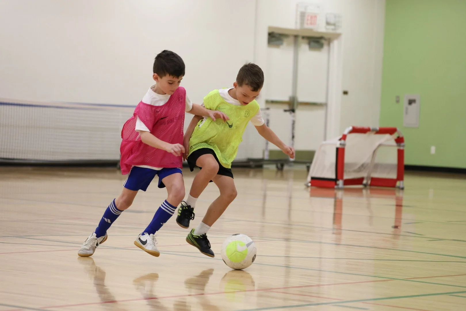 Two young boys playing indoor soccer, chasing a ball on a gym floor, one wearing a pink mesh jersey and blue shorts, the other in a yellow jersey and black shorts.