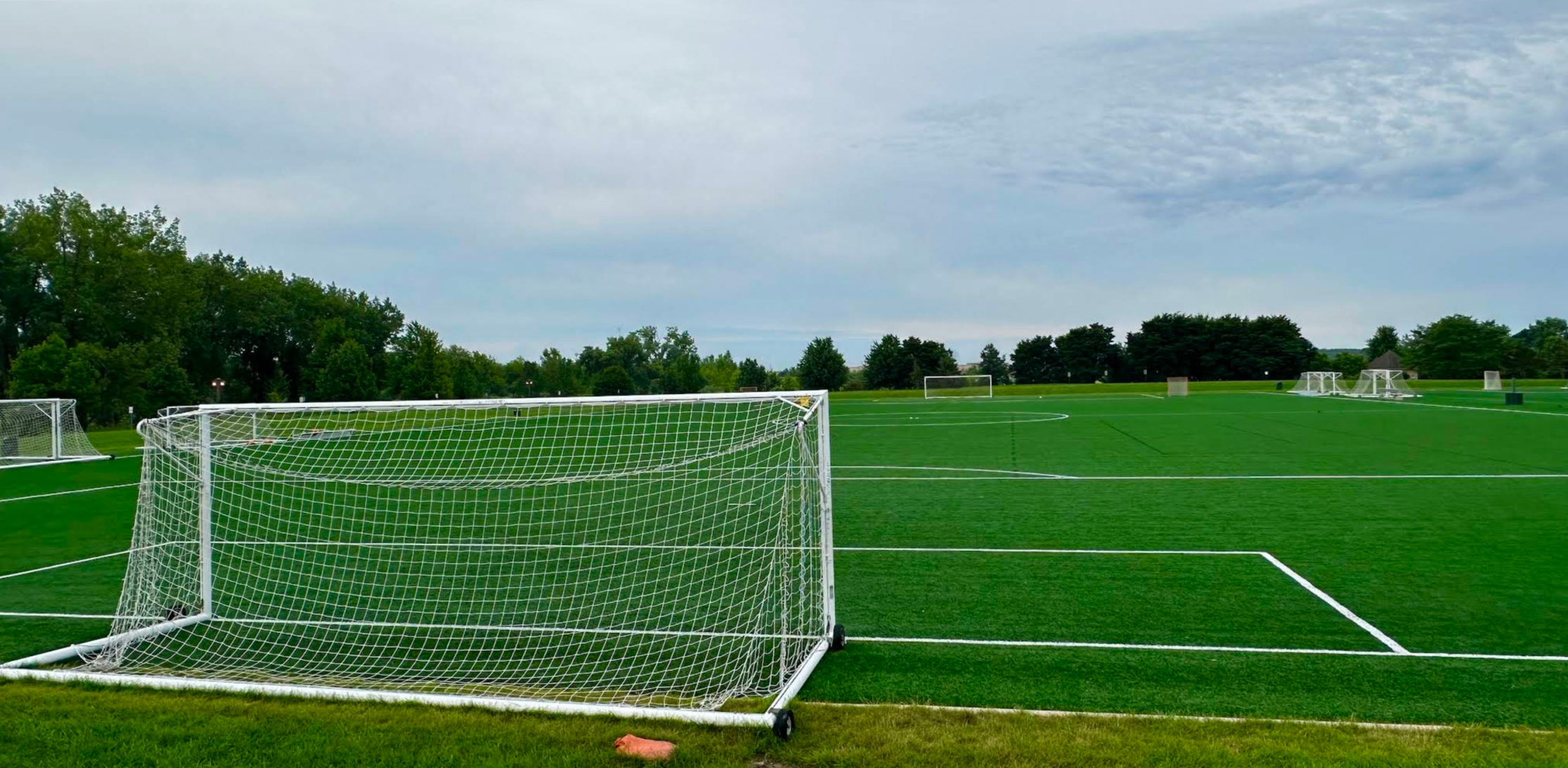 Empty soccer field with multiple goals, green grass, and cloudy sky.