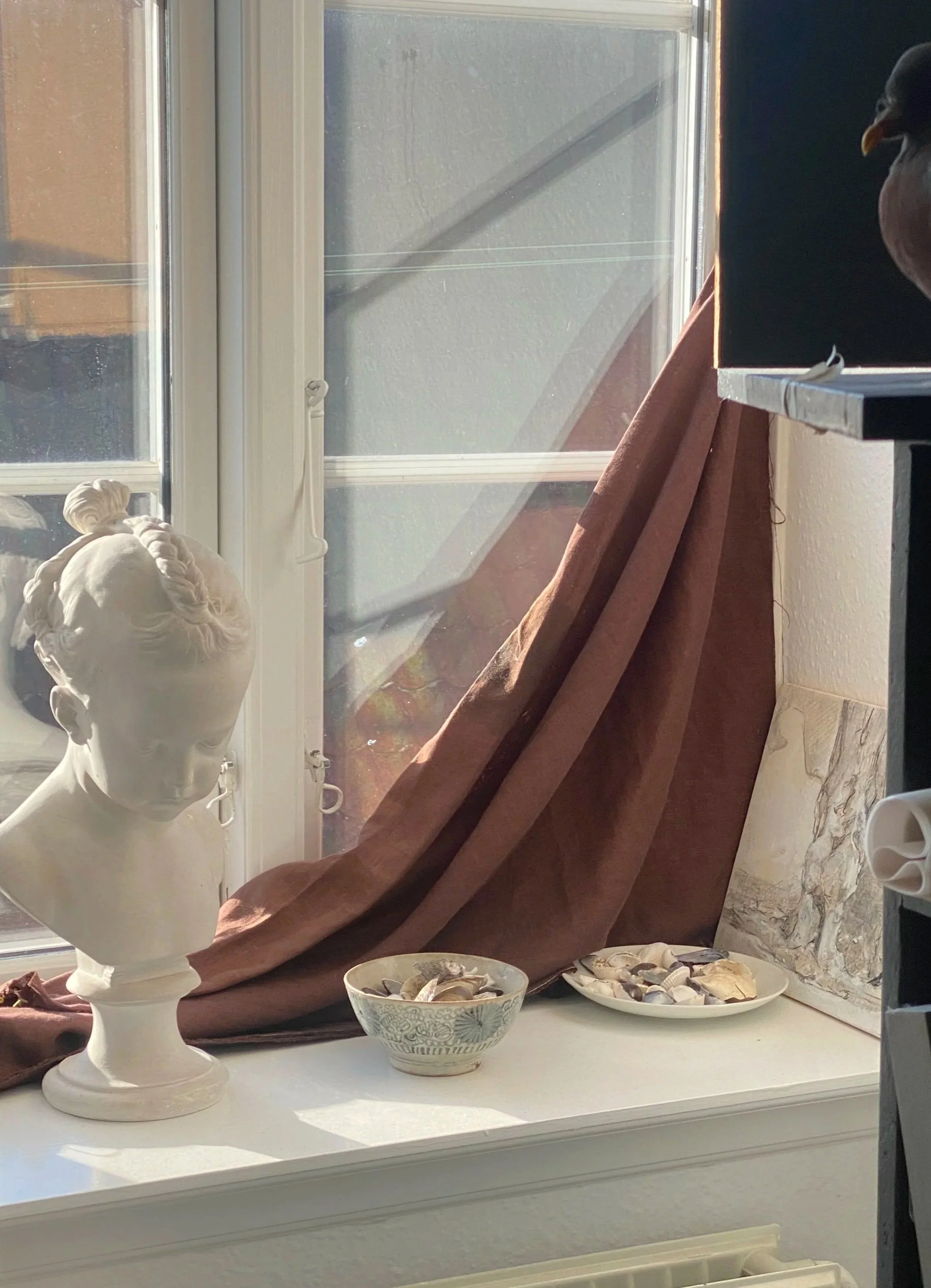 A white bust sculpture of a woman with braided hair, placed on a windowsill with brown and pink drapes, two bowls filled with seashells, and sunlight coming through a nearby window.