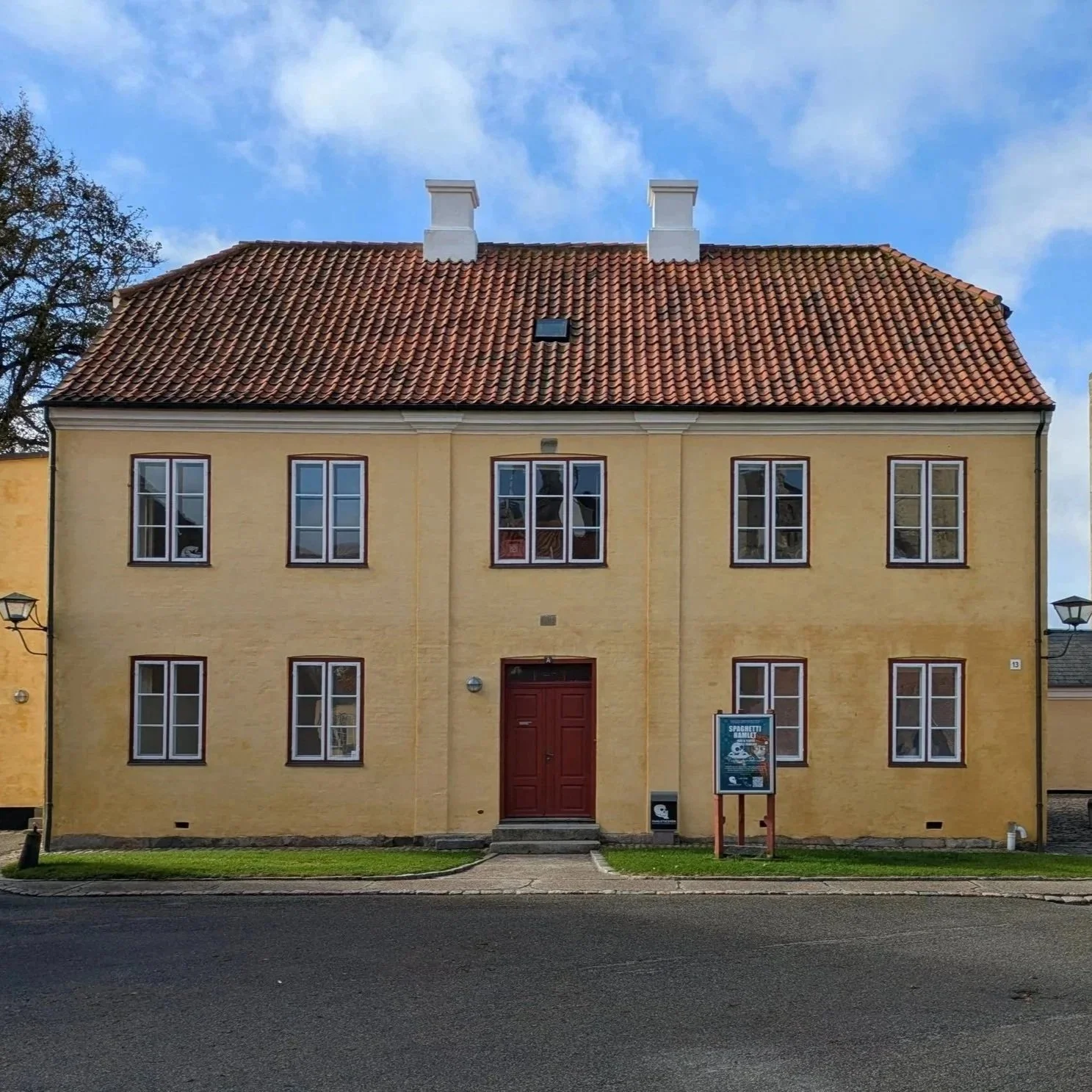 A yellow, three-story house with a red tiled roof and white chimneys, set against a partly cloudy sky. The house has six rectangular windows with white frames, a red front door, and a small sidewalk leading to the entrance. There is a signboard on the right side of the house and a grass lawn in front.