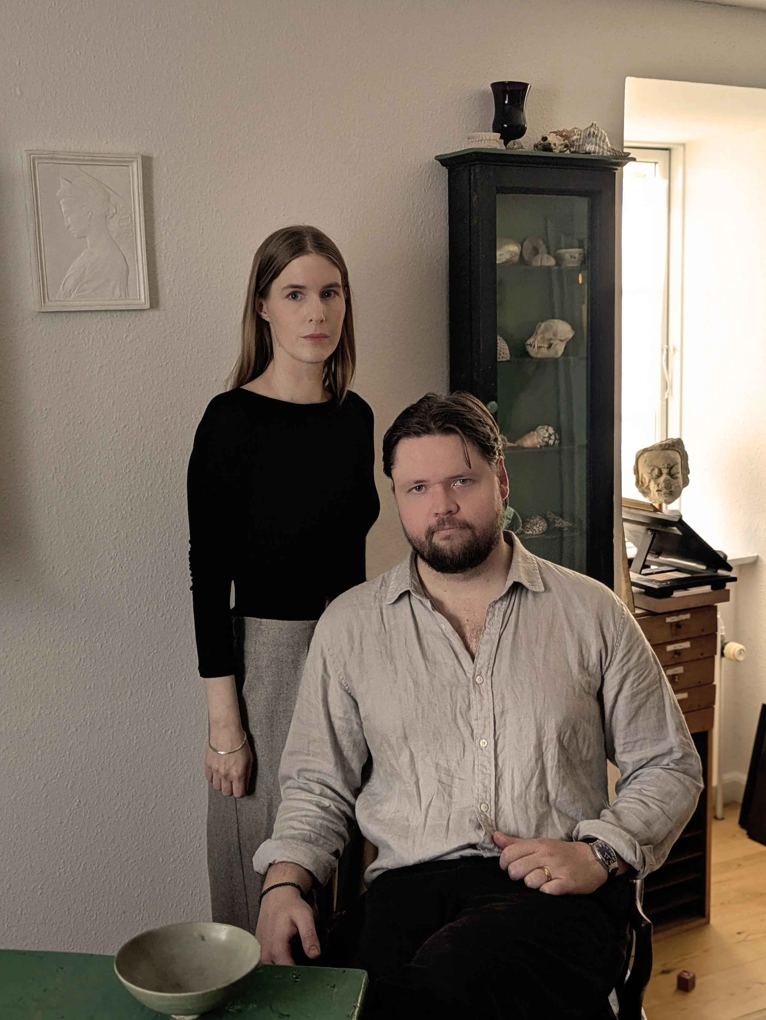 A woman and a man in a room with beige walls and wooden floors. The woman is standing with long brown hair, wearing a black top and gray pants. The man is seated, has dark hair and a beard, and is wearing a light-colored, slightly wrinkled shirt. There is a dark green cabinet with glass doors behind them, containing multiple shells and a skull. To the right, there is a window letting in natural light, and on the left wall, there’s a framed profile silhouette of a woman. A green table with a bowl on it is also visible in the foreground.