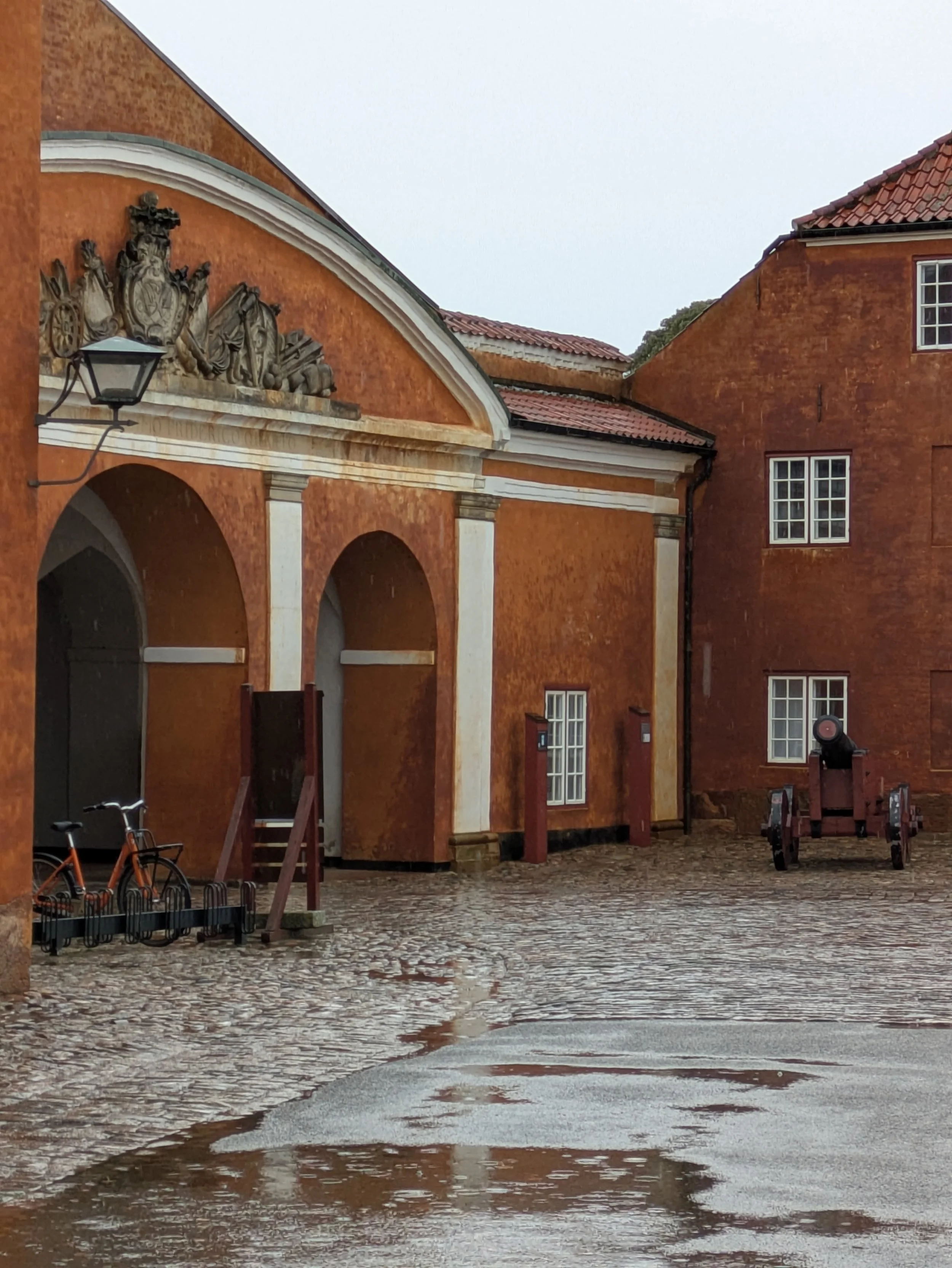A historic orange brick building with white accents, arched doorway, cobblestone courtyard and old cannon.