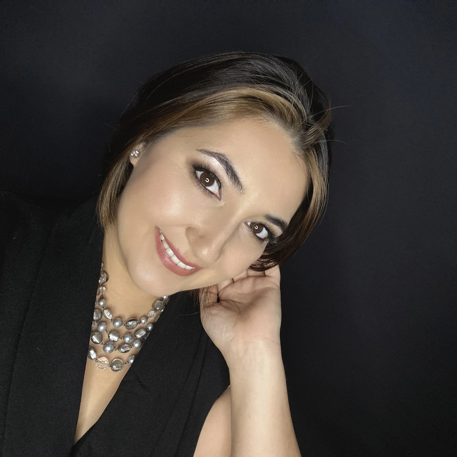 Close-up portrait of a smiling woman with short brown hair, wearing pearl jewelry and a black top, posing against a dark background.