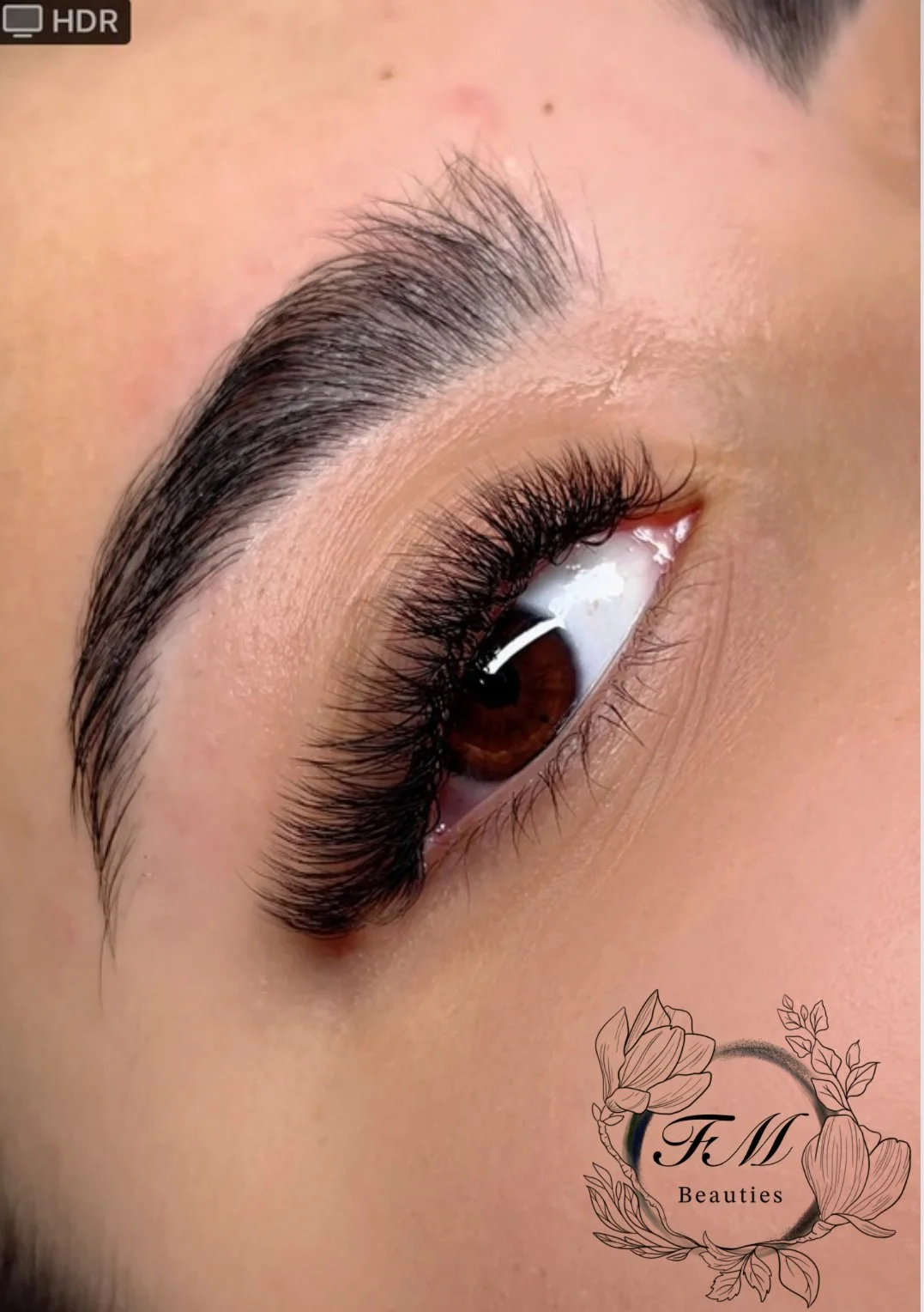 Close-up of a well-groomed eyebrow and long, dark eyelashes on a person with brown eye and natural skin. There is a logo with flowers and the text 'FtM Beauties' in the bottom right corner.