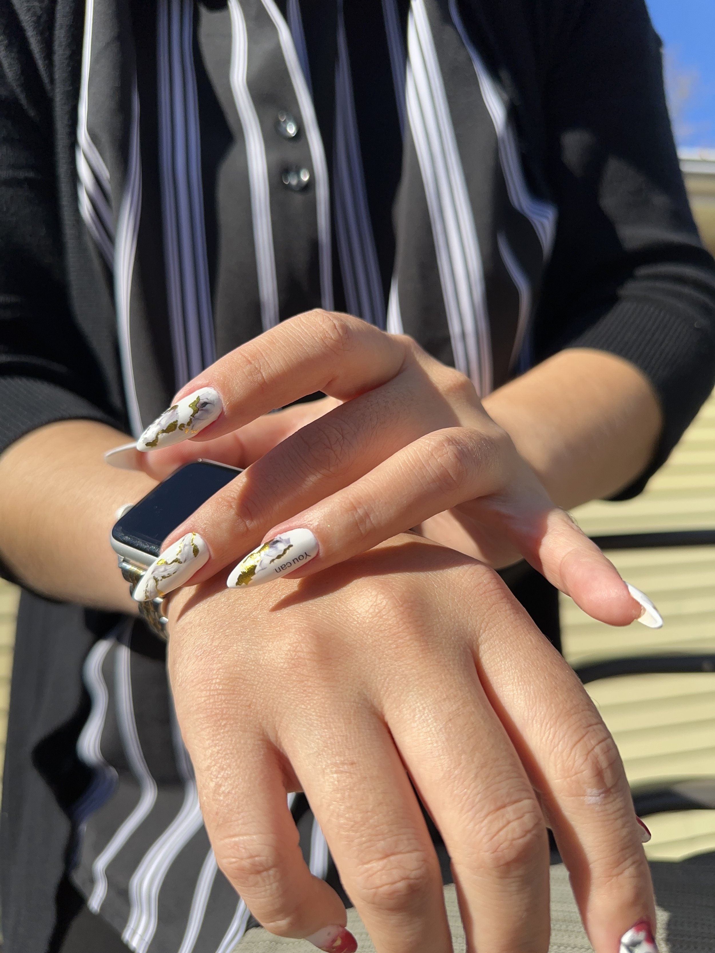 Close-up of two hands, one with a smartwatch and painted nails, overlapping. The background shows a person wearing a black shirt with vertical white and purple stripes.