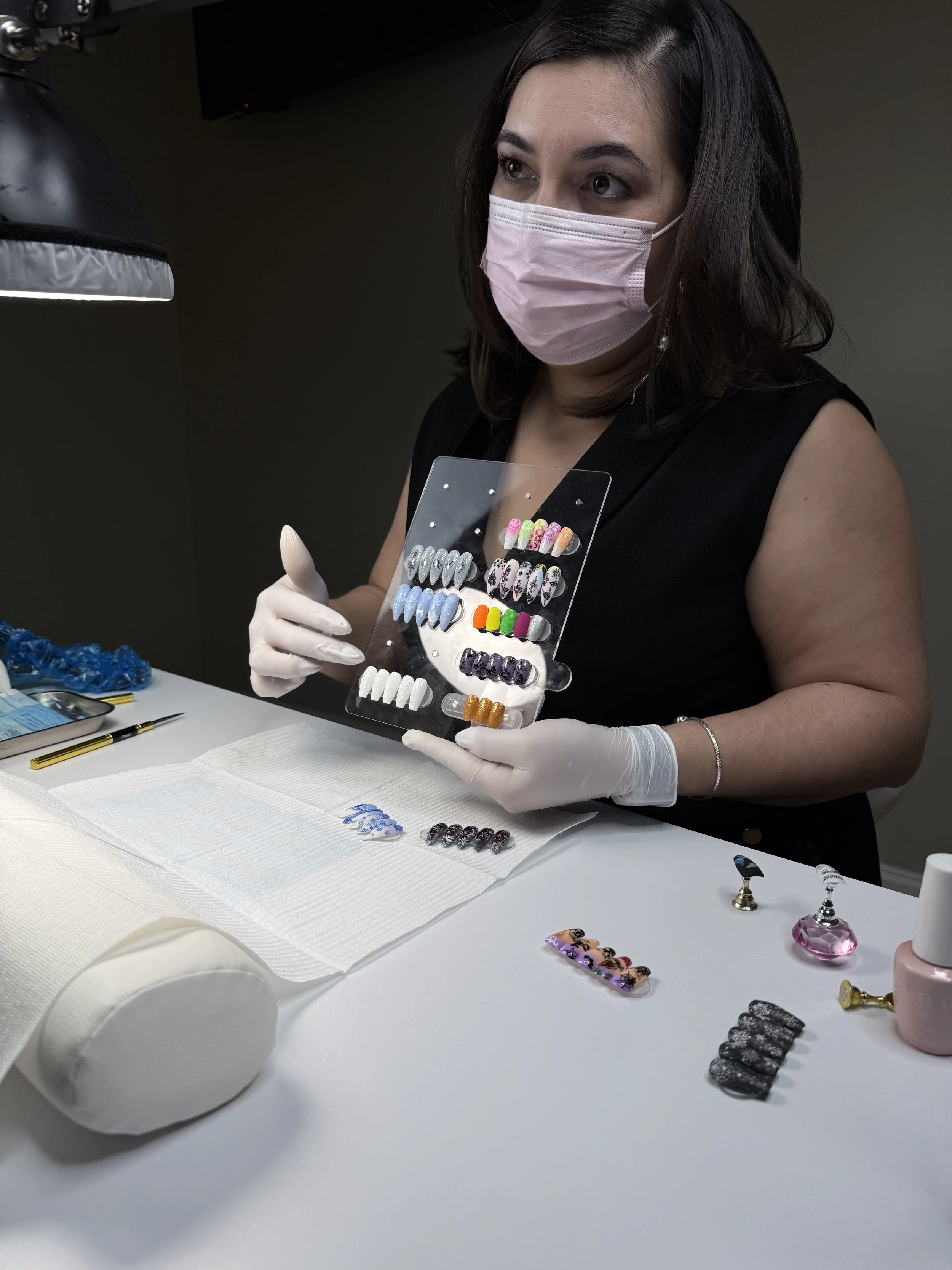 A woman wearing a pink face mask and white gloves holding a display of colorful artificial nails in a nail salon or studio.