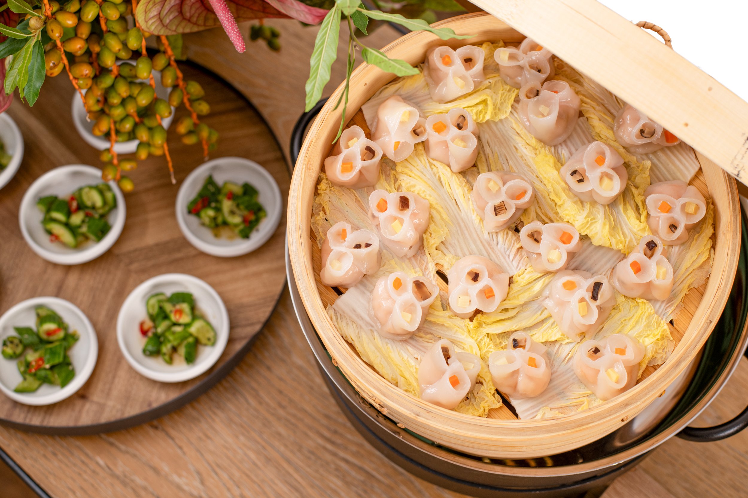 Steamer basket filled with raw dumplings on layers of napa cabbage, nearby small bowls with chopped vegetables, set on a wooden surface.