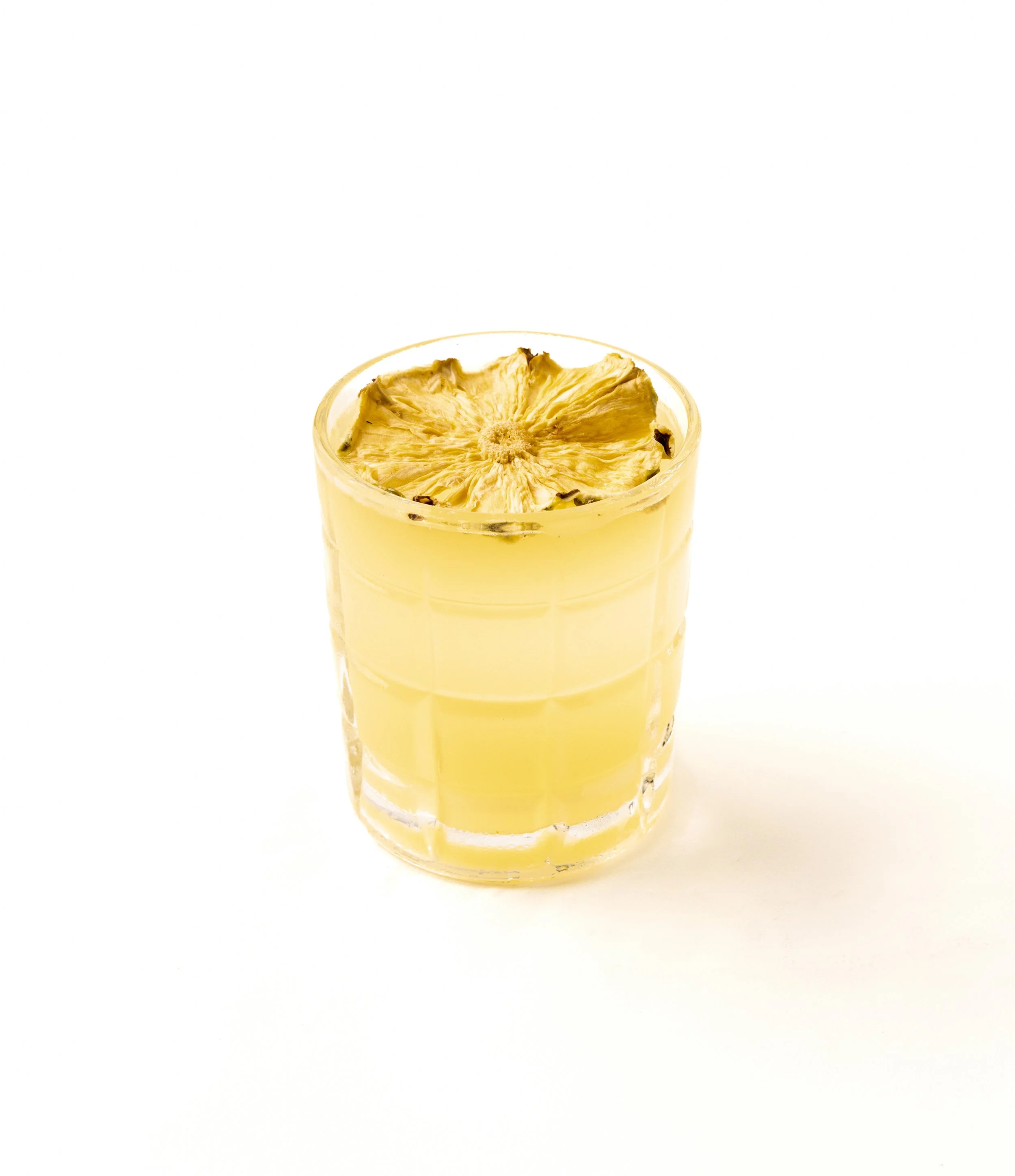A clear glass containing a yellow beverage with a dried pineapple slice on top, set against a white background.