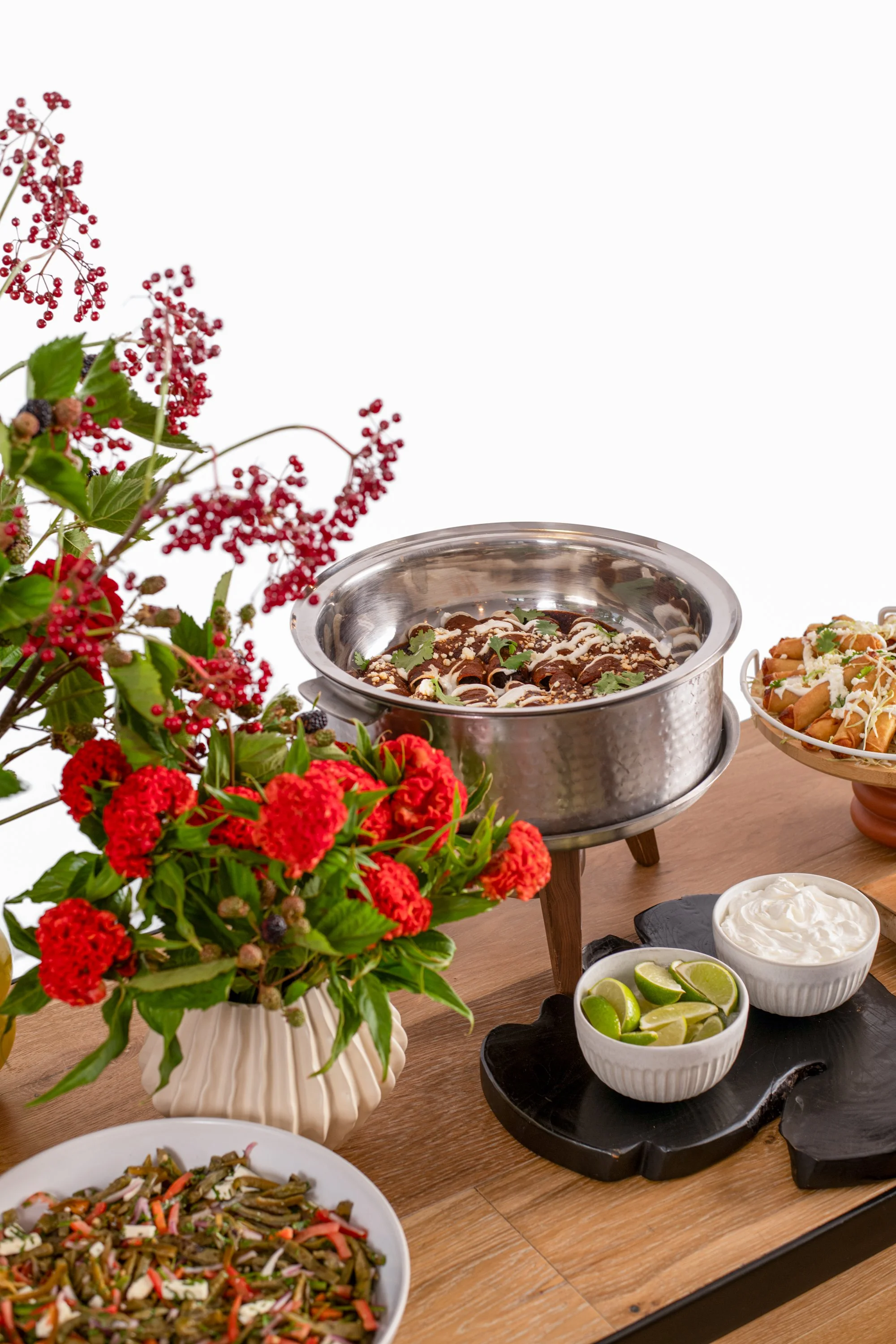 Mexican food spread with a large bowl of tamales, side dishes of lime wedges and sour cream, fresh flower arrangement, and other dishes on a wooden table.