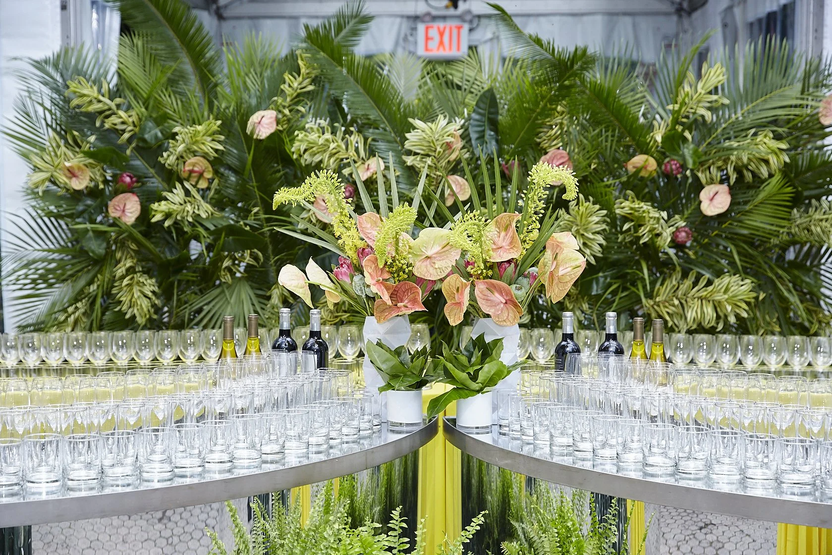 Elegant floral arrangement of pink and green flowers in white vases, surrounded by rows of empty glasses and bottles on a table, with lush green plants in the background and an exit sign overhead.