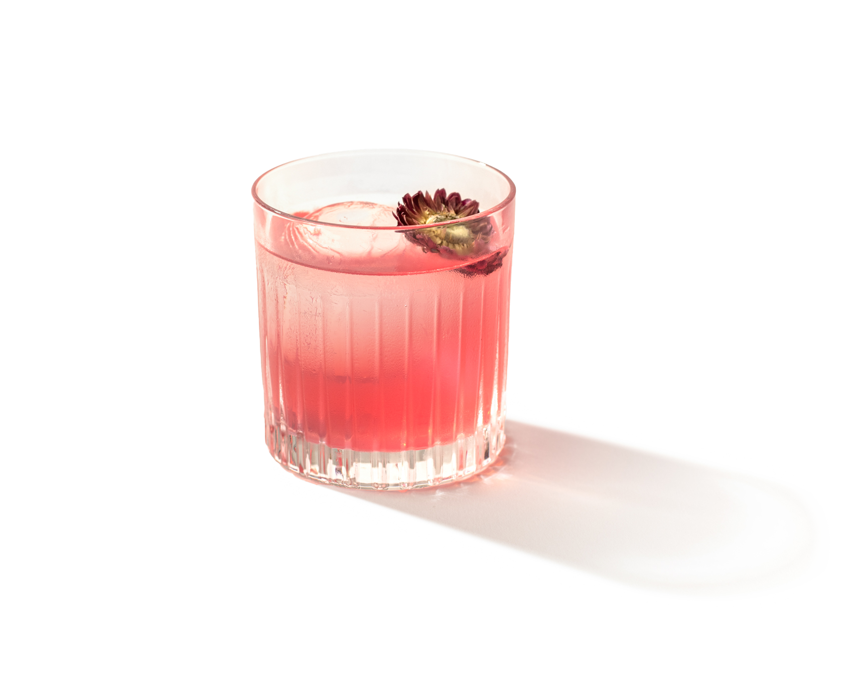 Pink cocktail in a glass with a floral garnish, casting a shadow on a white background.