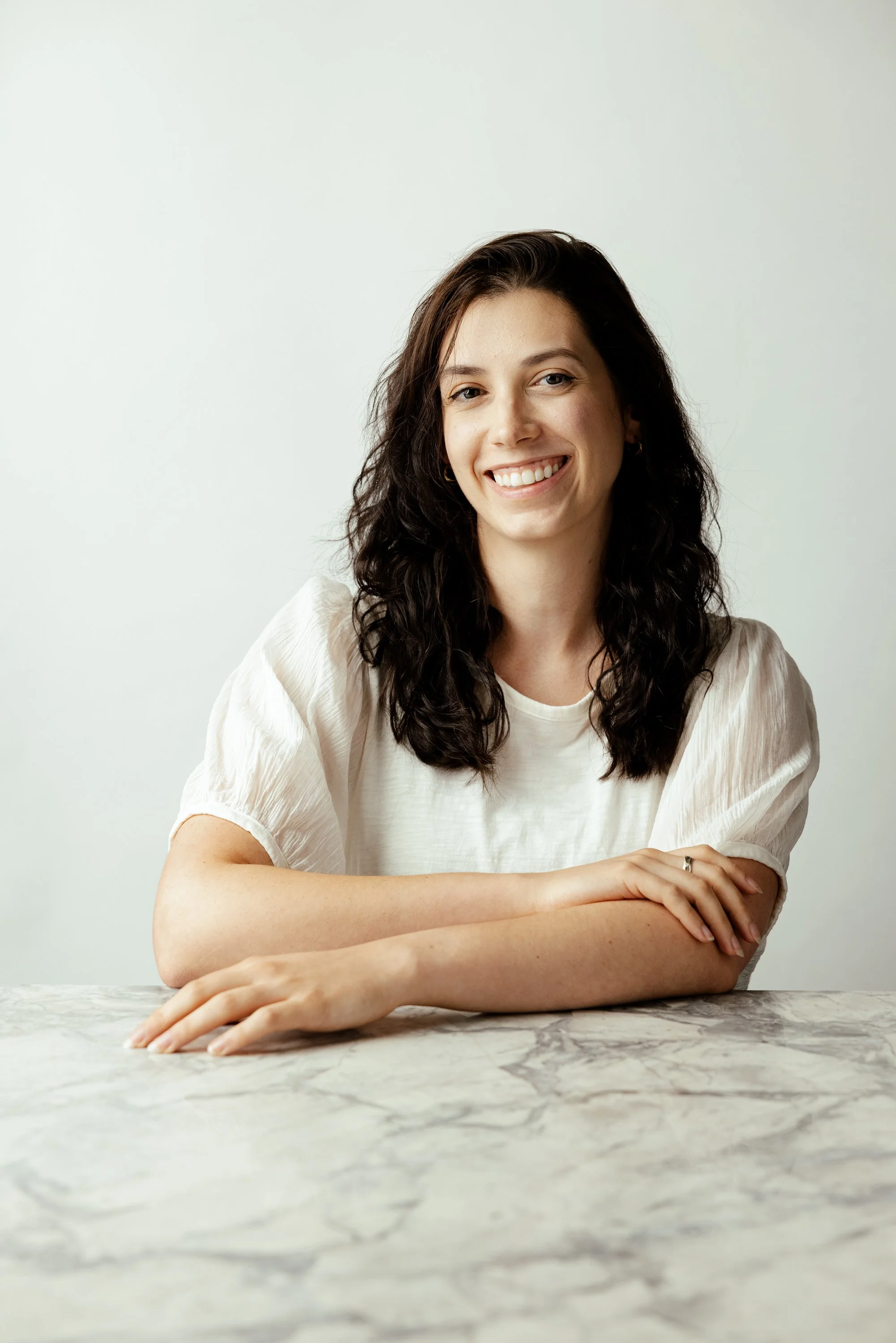 A young woman with dark, curly hair smiling, sitting with her arms crossed on a marble table against a plain light-colored background.
