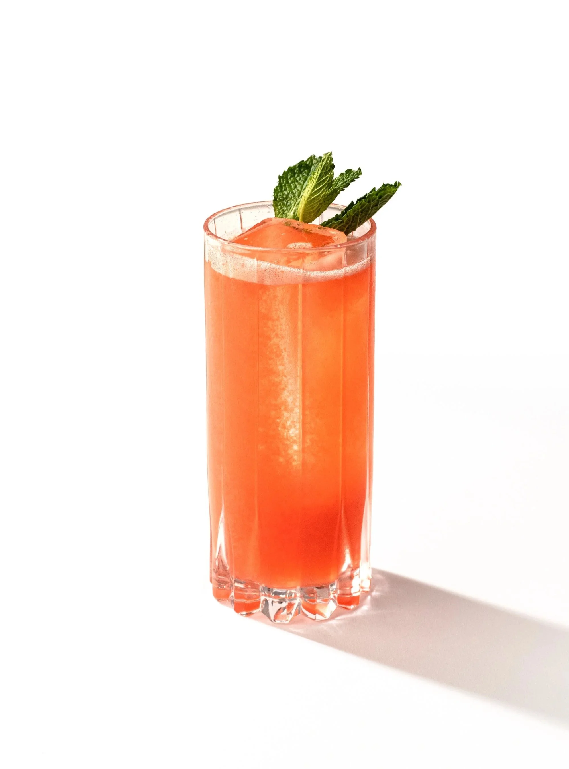 A tall glass of pink cocktail garnished with fresh mint leaves, set on a white surface with a shadow.