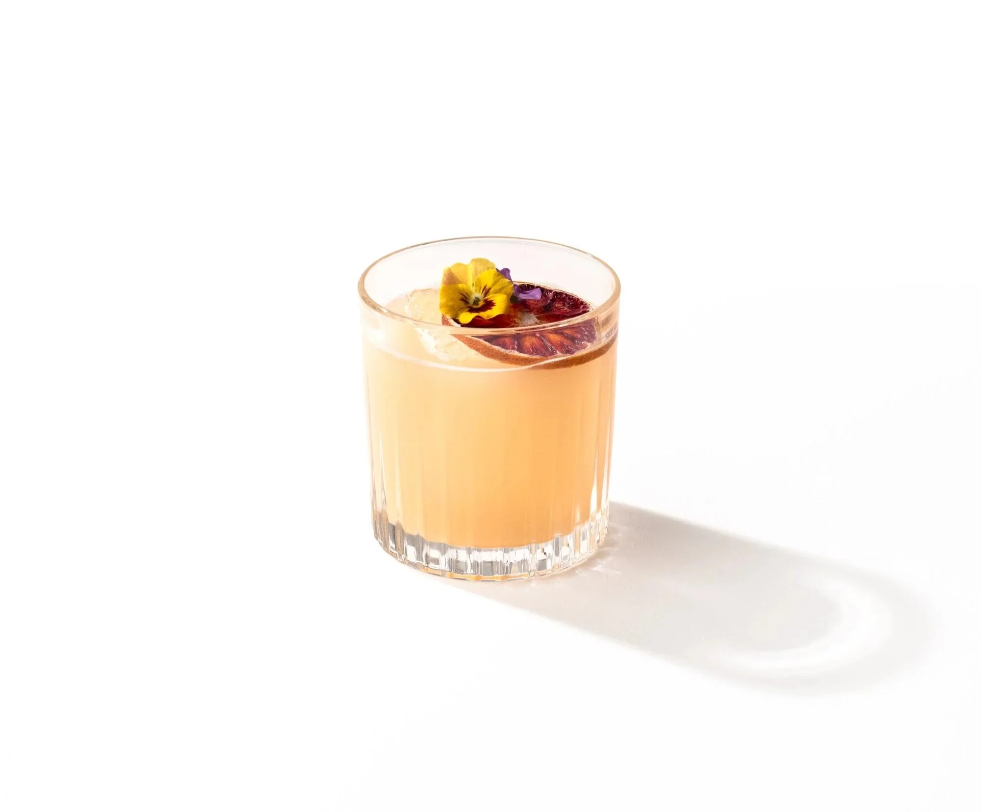 A clear glass of pink cocktail garnished with a purple pansy flower and a slice of blood orange on a white background.