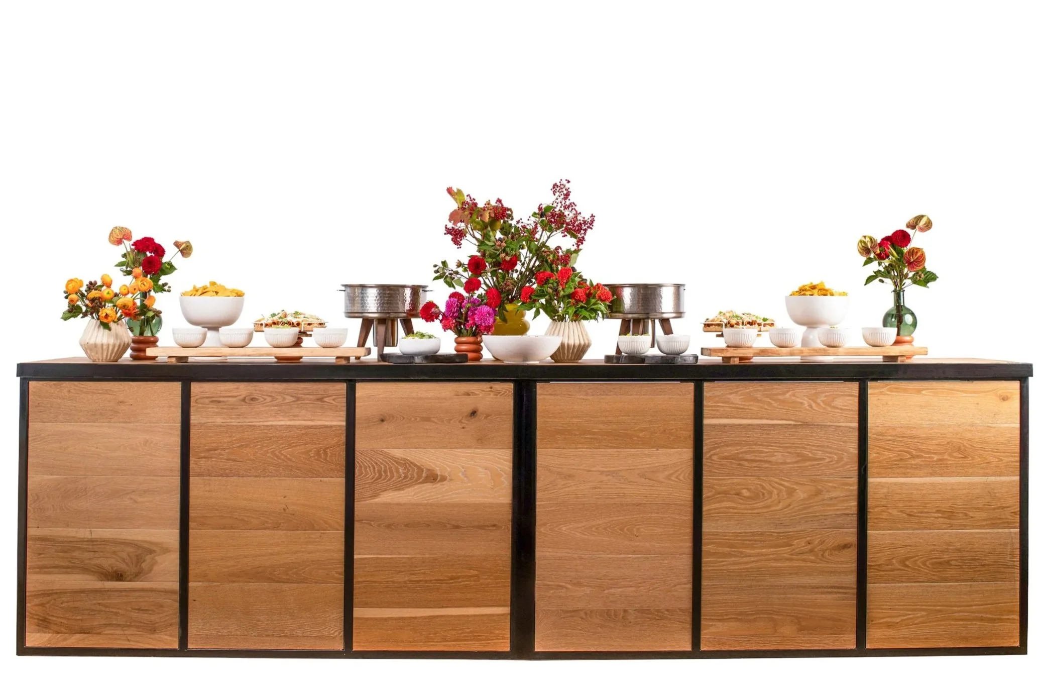 A buffet table decorated with colorful flowers, bowls of food, and serving dishes against a white background.