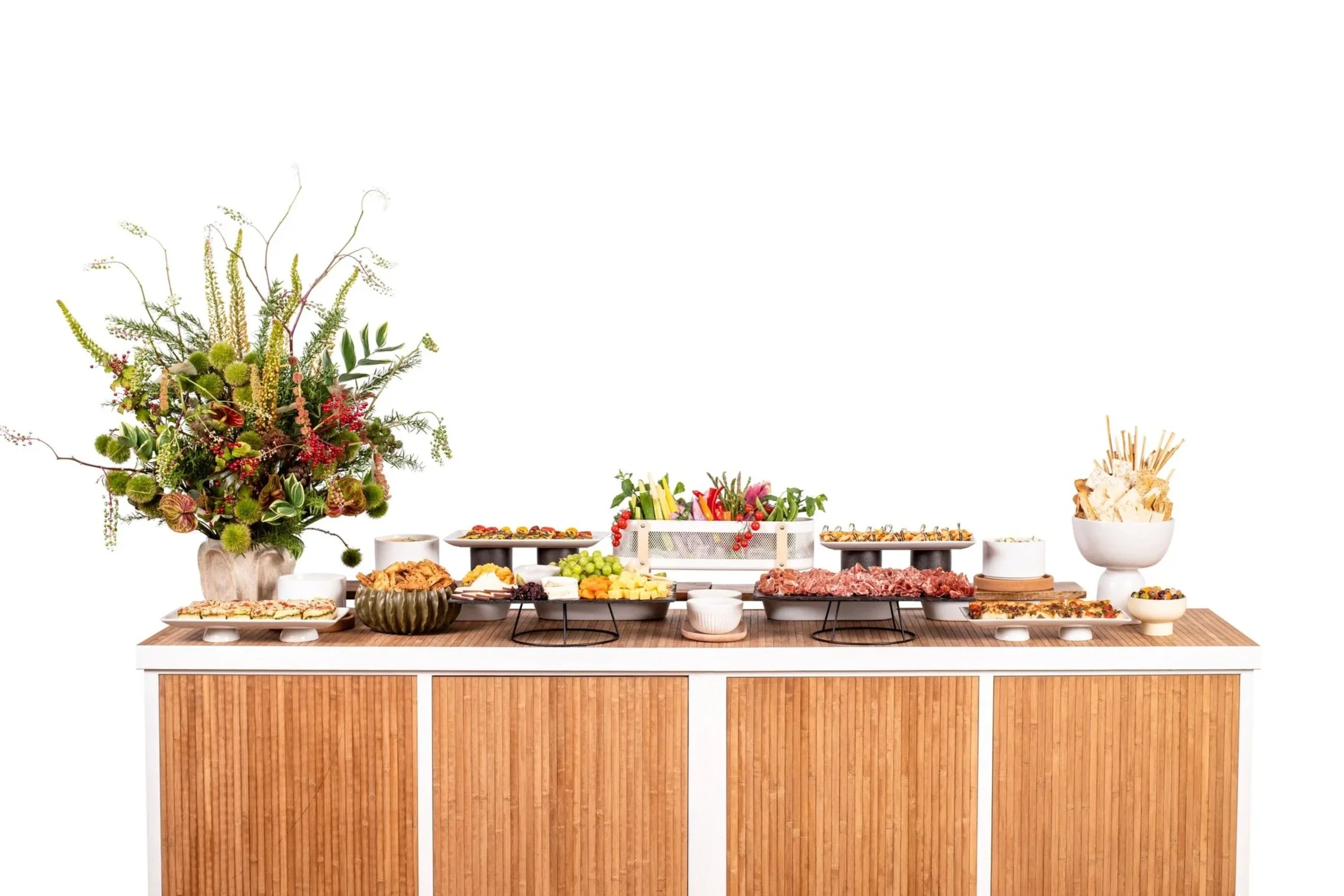 A buffet table with a large floral arrangement on the left and various dishes including fruits, meats, chips, and dips