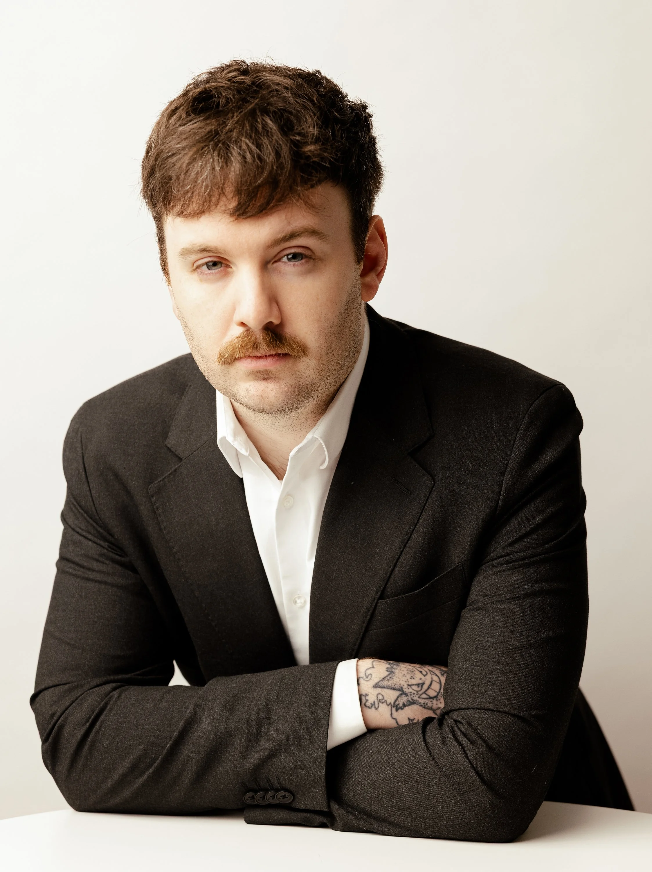 A man with brown hair, a mustache, and a serious expression, wearing a black suit and white shirt, sitting at a white table with his arms crossed, against a plain white background.