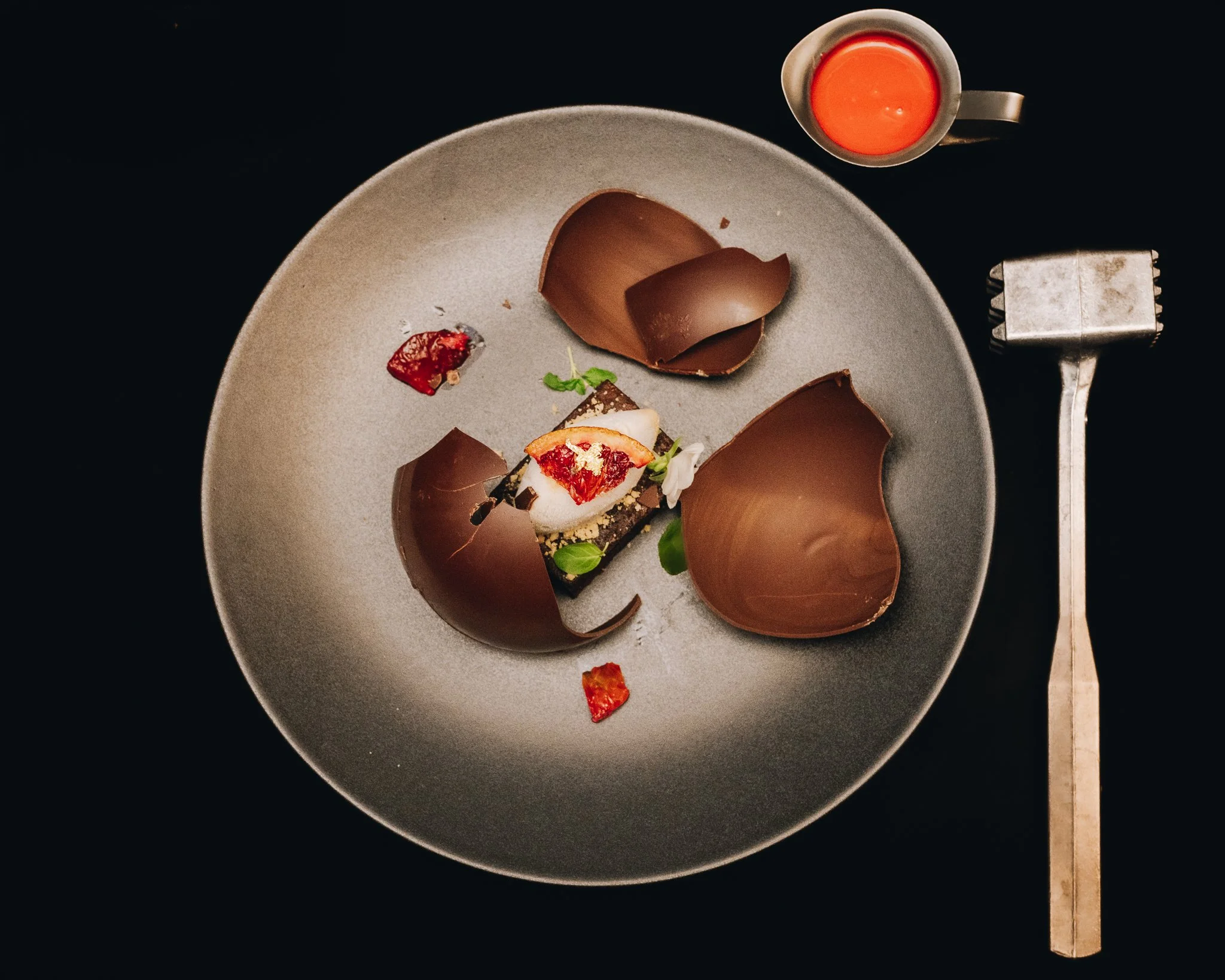 A plate with a dessert garnished with strawberries, mint, and chocolate shavings, three large chocolate curls, a small cup of orange sauce on the side, and a silver meat tenderizer tool on a dark background.