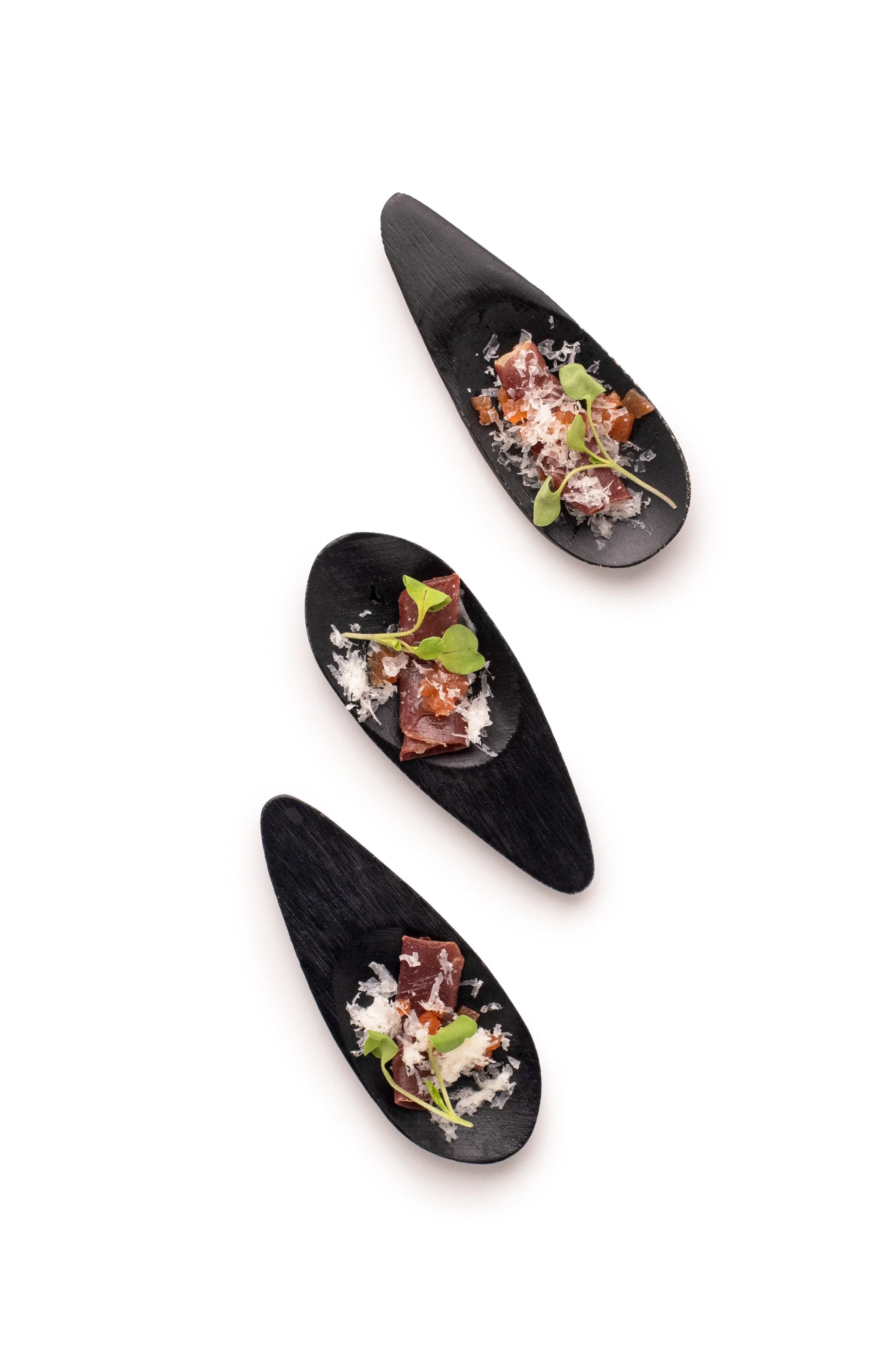 Three black spoons with small portions of gourmet food, garnished with microgreens and grated cheese, arranged vertically on white background.