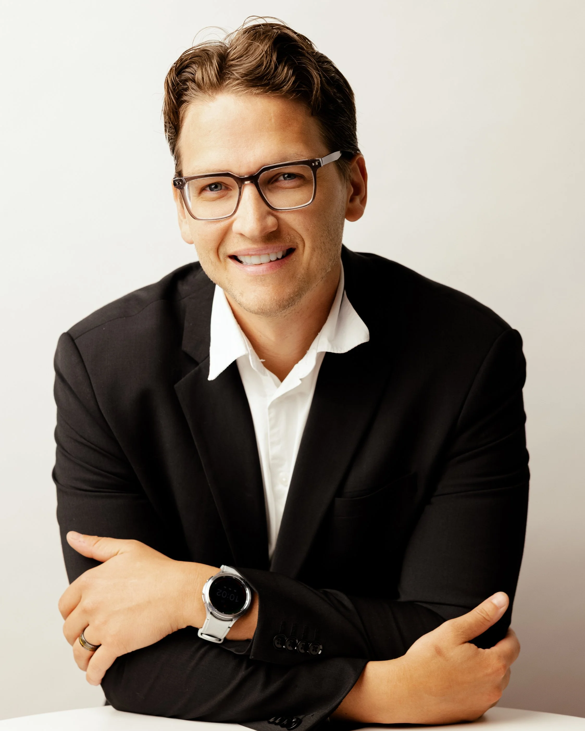 A smiling man with glasses in a black suit and white shirt, wearing a smartwatch, arms crossed, against a plain white background.