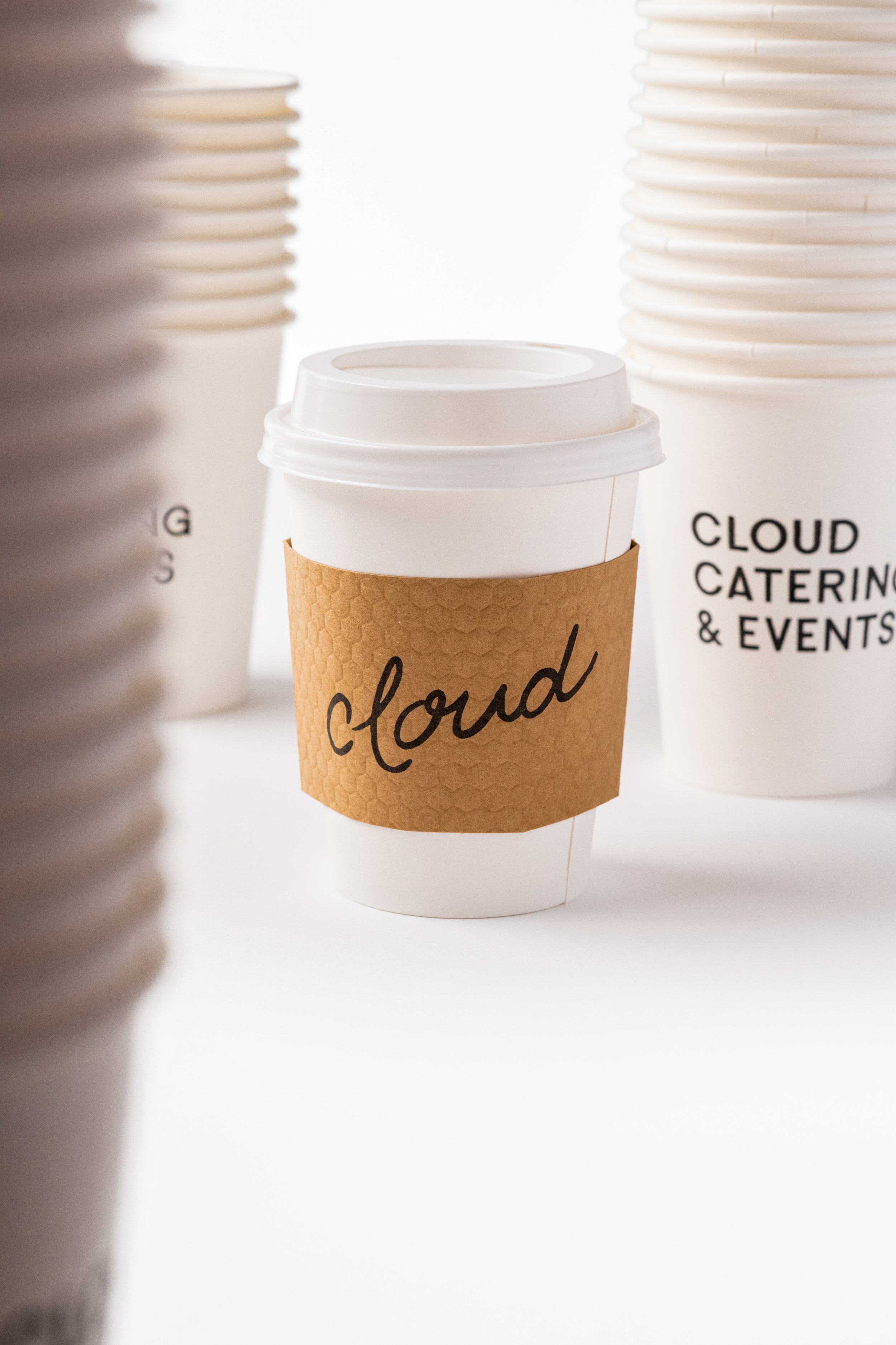 A white disposable coffee cup with a brown sleeve labeled 'cloud', surrounded by stacks of similar cups with text 'CLOUD CATERING & EVENTS' and partially visible cups around.