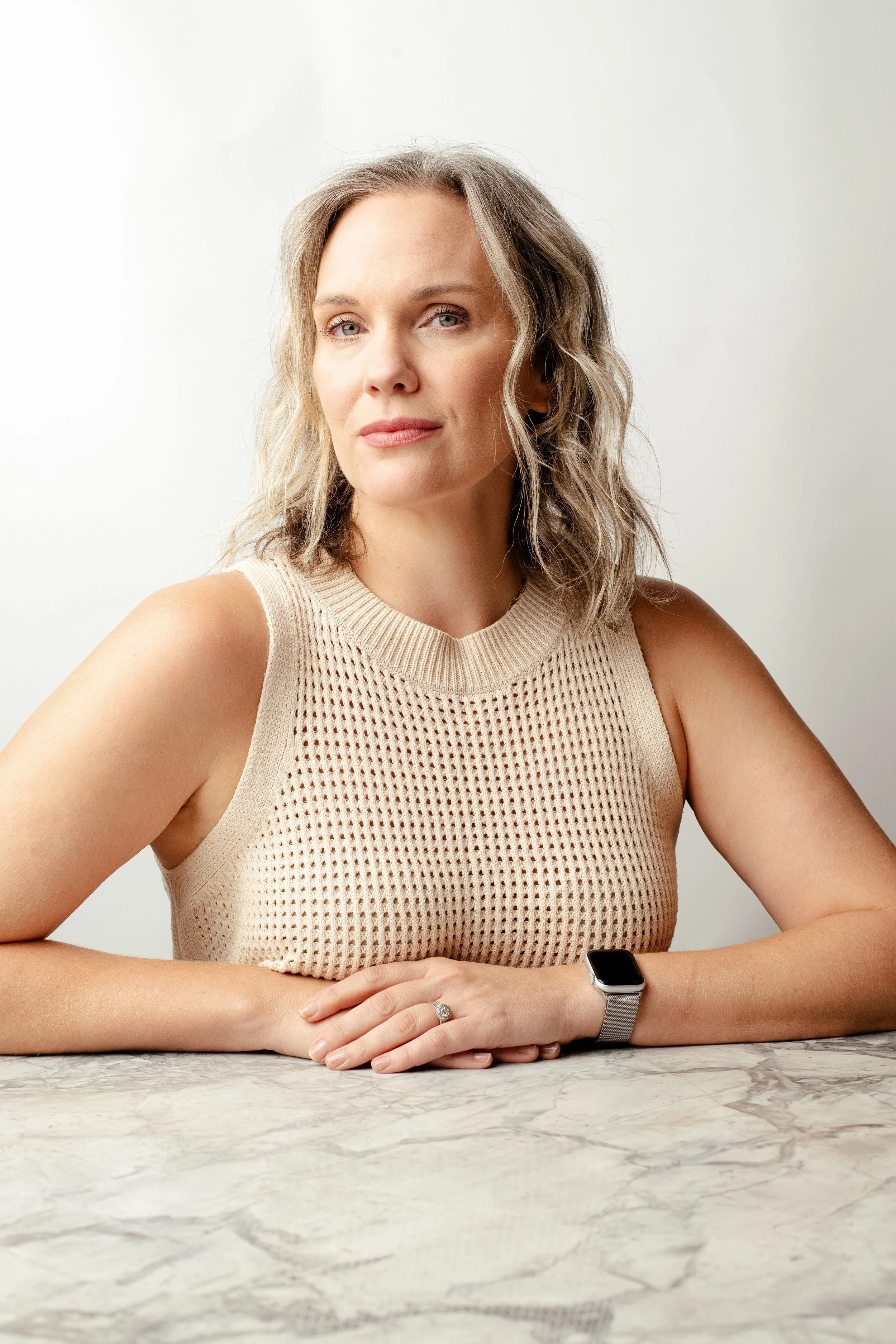 A woman with shoulder-length wavy blonde hair and blue eyes sitting at a marble table, wearing a sleeveless beige knit top and a smartwatch.