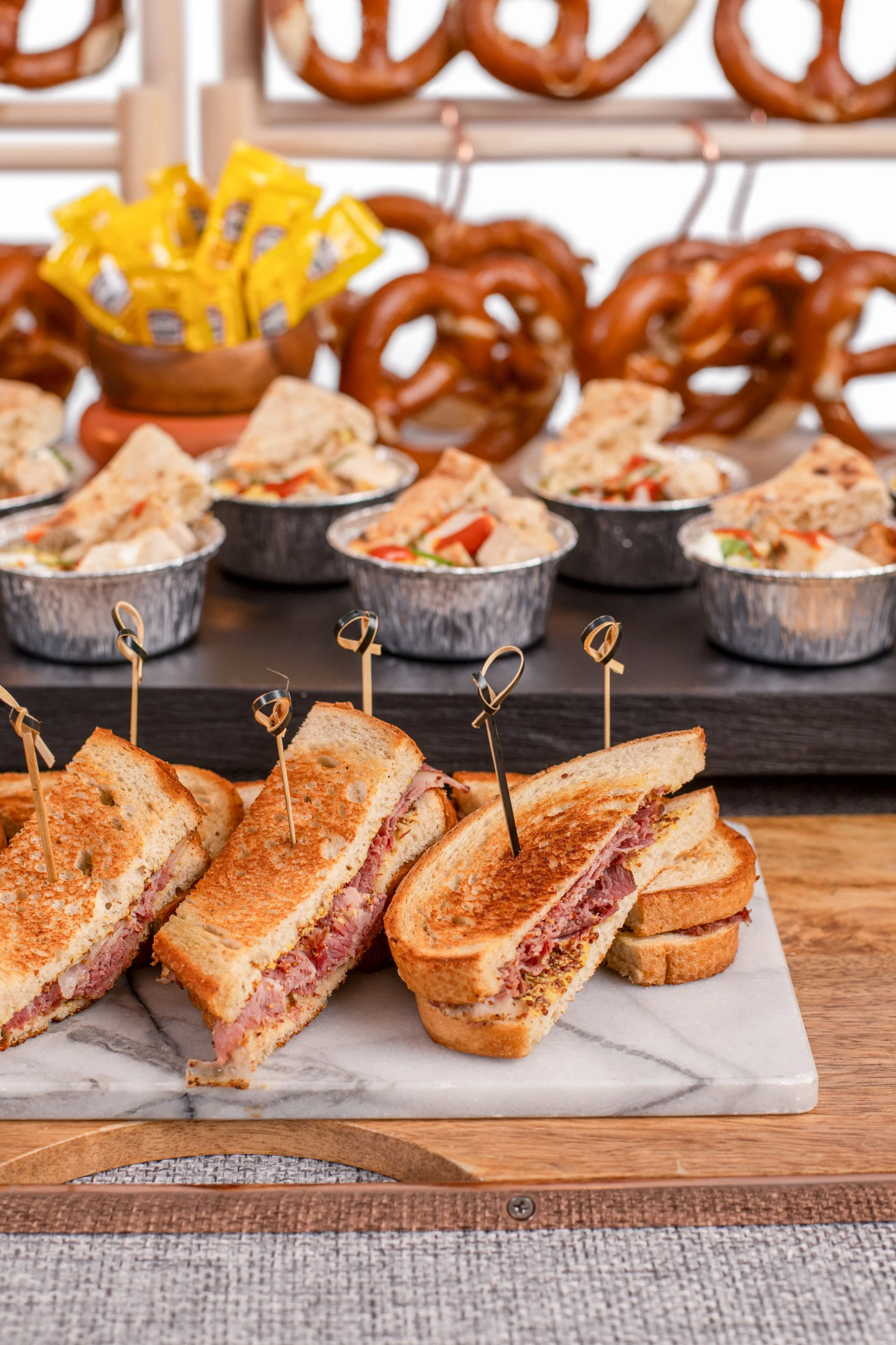 Close-up of ham and cheese sandwiches with skewers, assorted finger foods, pretzels, and cheese packets on a buffet table.