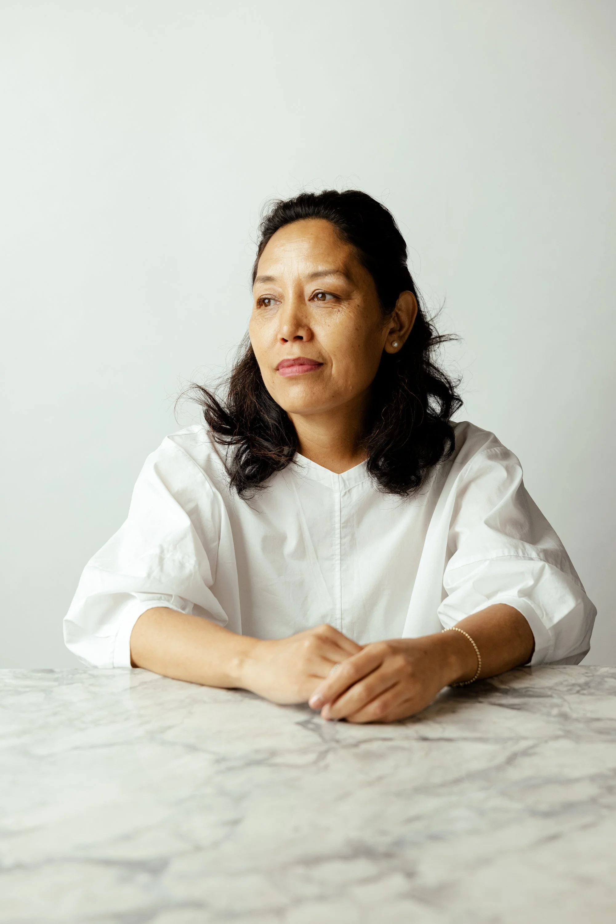A woman with dark hair, wearing a white top, sitting at a marble table, looking thoughtful against a plain white background.
