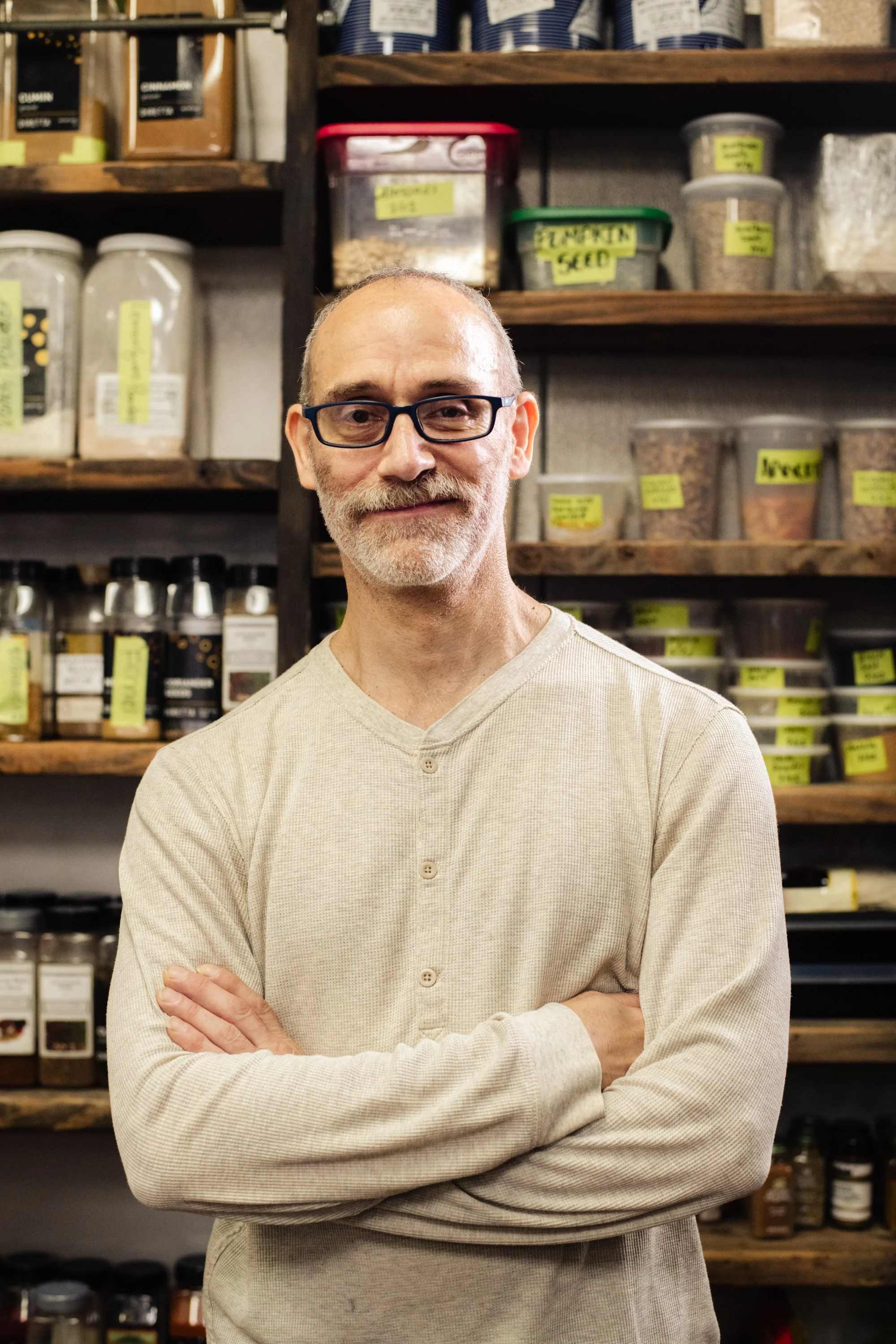 A man with glasses and a beard standing with arms crossed in front of shelves filled with labeled jars and containers of herbs and spices in a store or pantry.