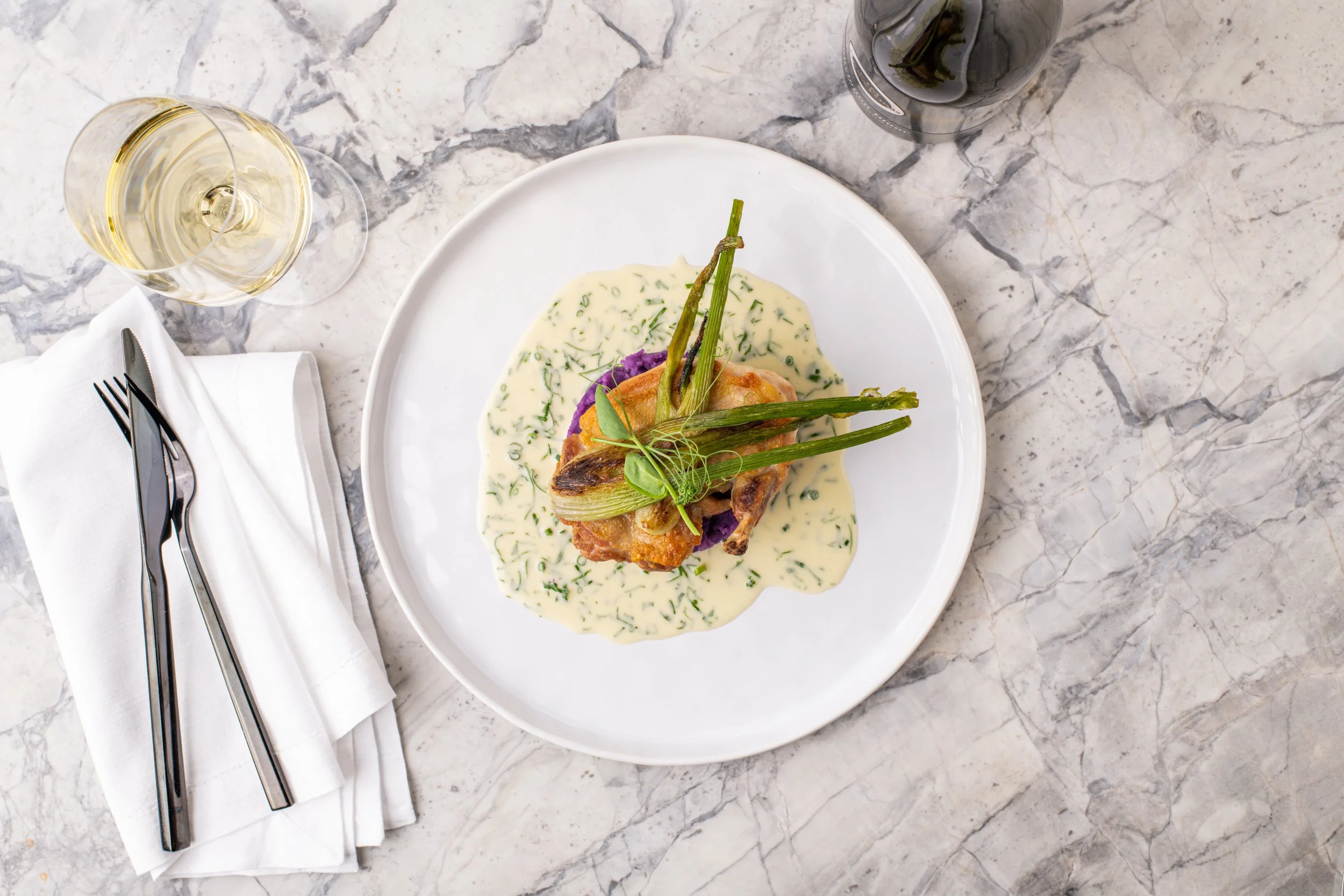 A plate of roasted chicken with grilled vegetables and creamy herb sauce, accompanied by a glass of white wine and a bottle of white wine on a marble table.