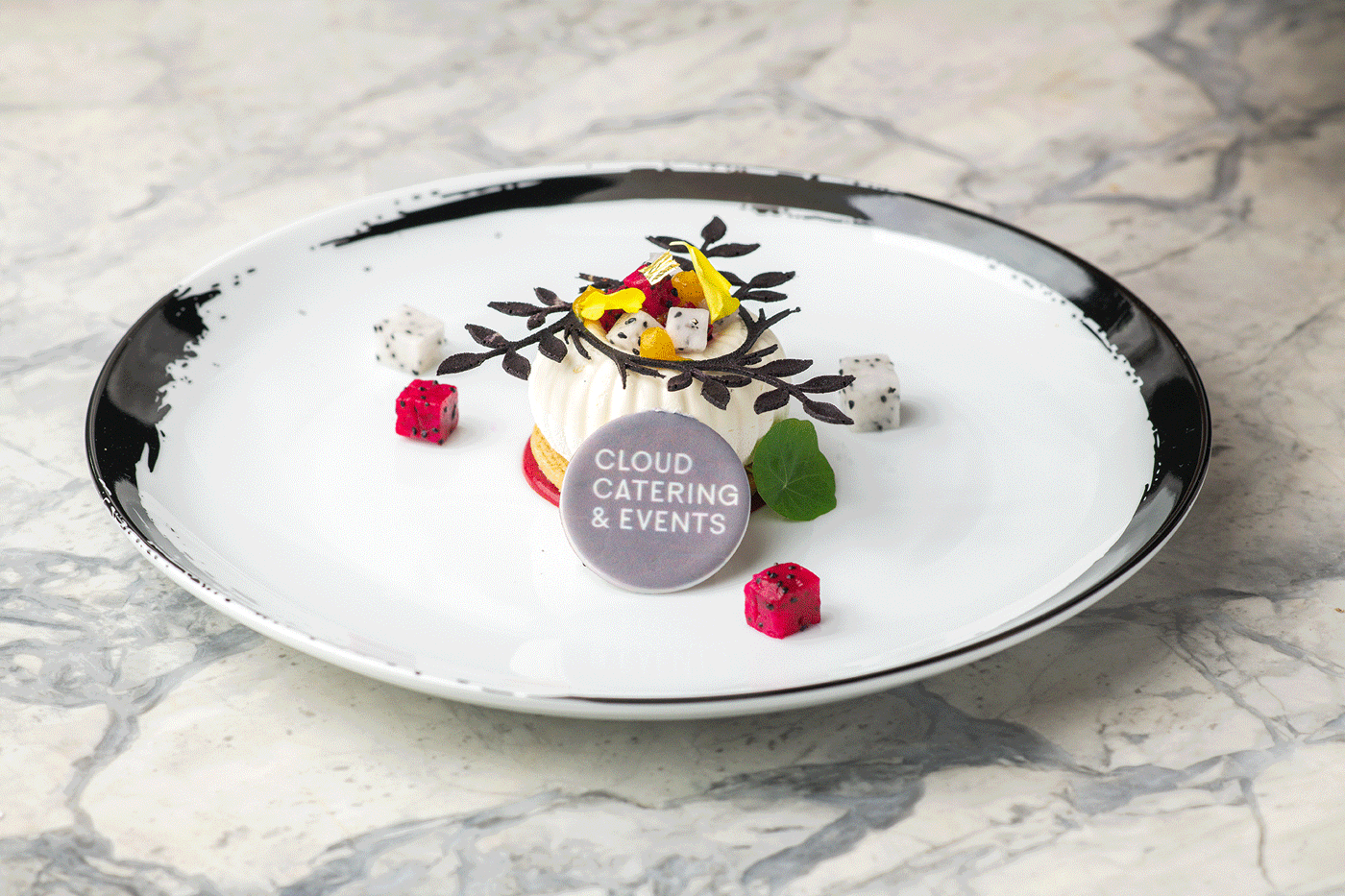 Elegant dessert with white mousse topped with chocolate decorations, fruit pieces, and edible flowers, served on a black and white plate with a label that reads 'CLOUD CATERING & EVENTS.'