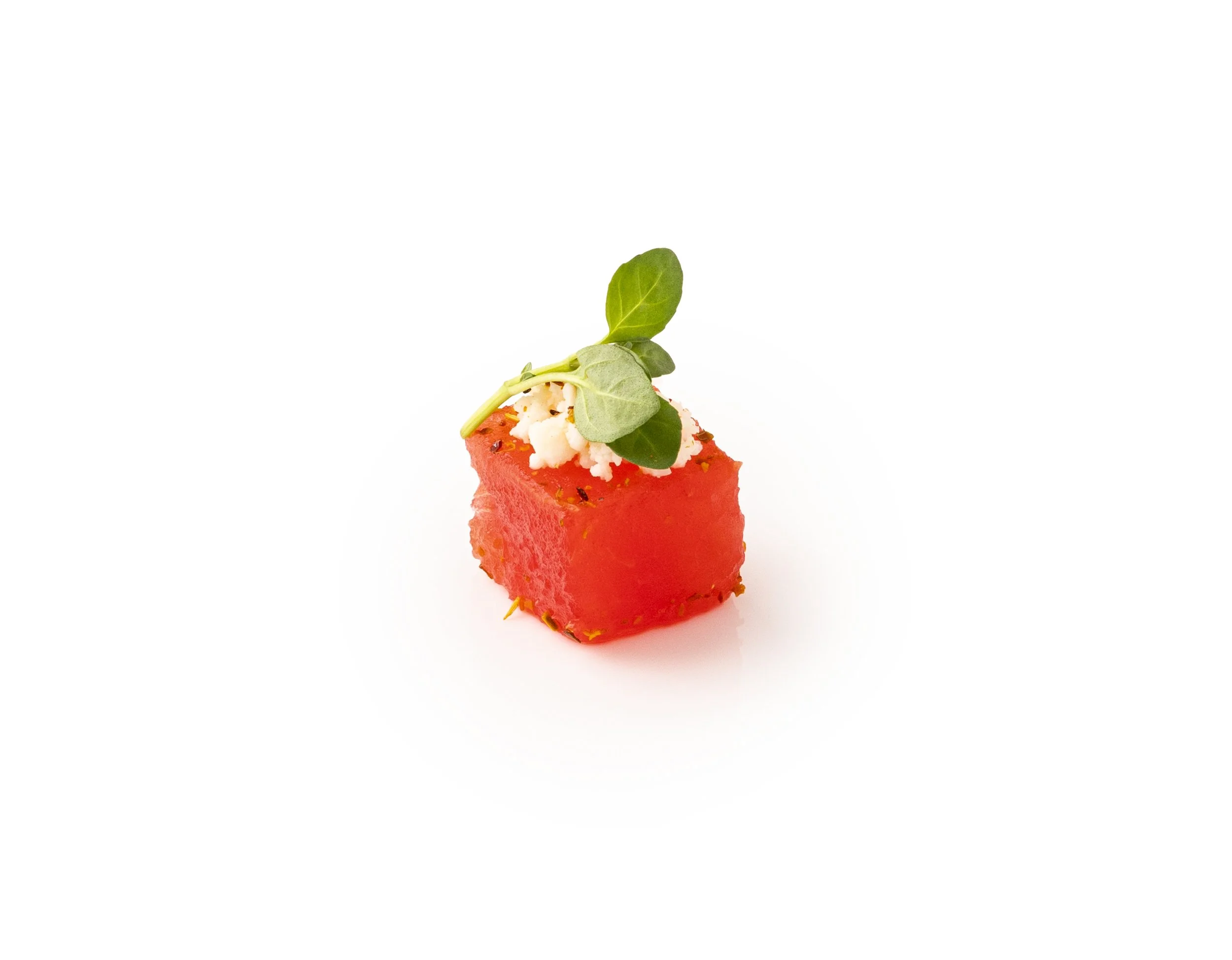 A small square piece of watermelon garnished with microgreens and some cheese, placed on a white background.