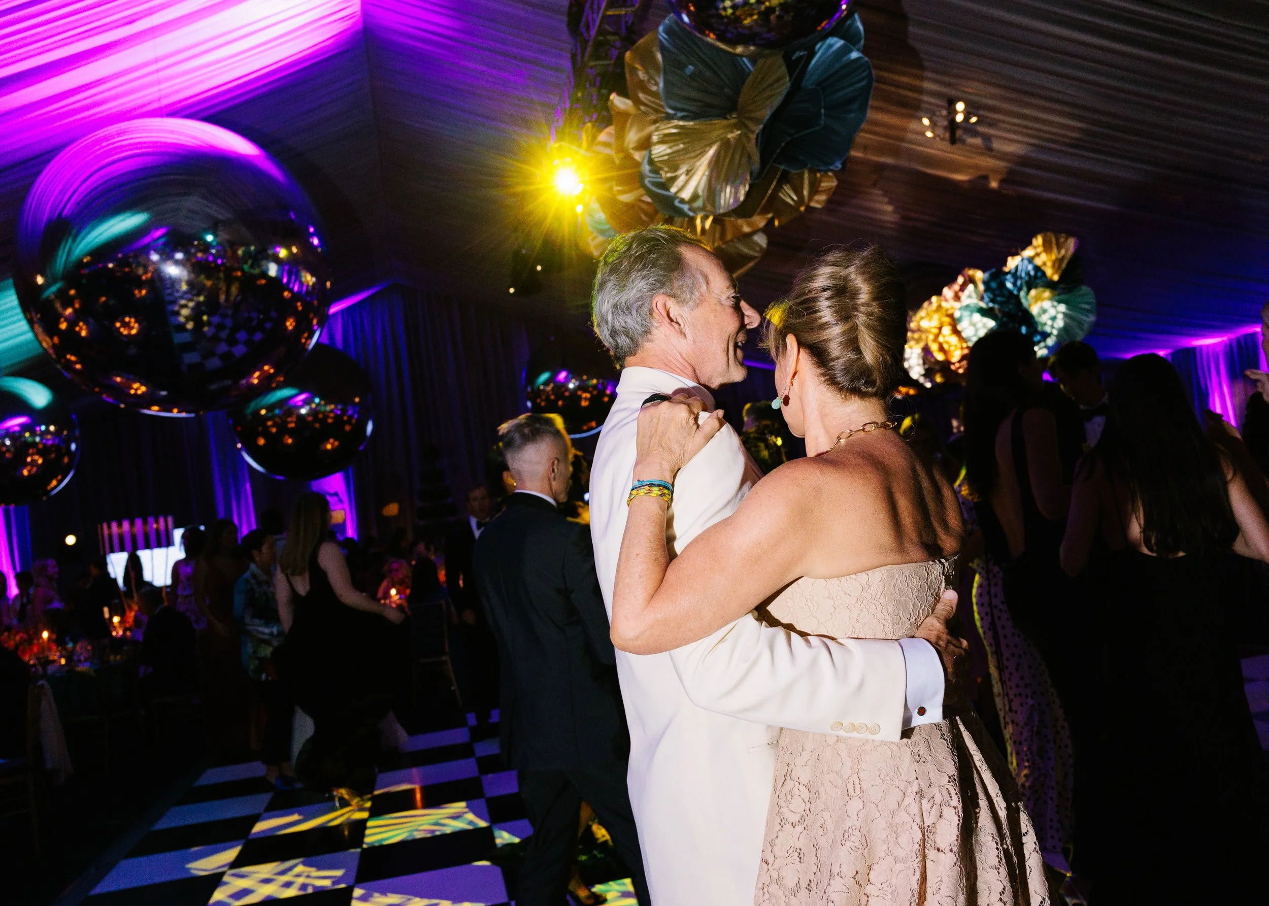 An elderly man and woman dancing closely at a party or wedding, surrounded by other guests in a decorated venue with purple lighting and large hanging balloons.