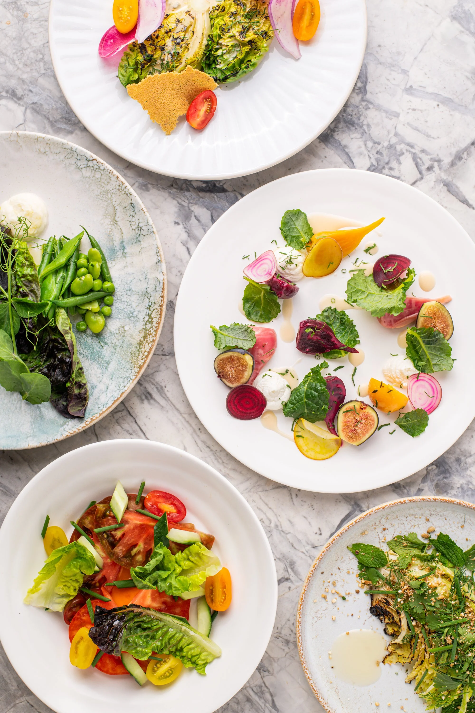 Assorted colorful salads on white plates on a marble table.
