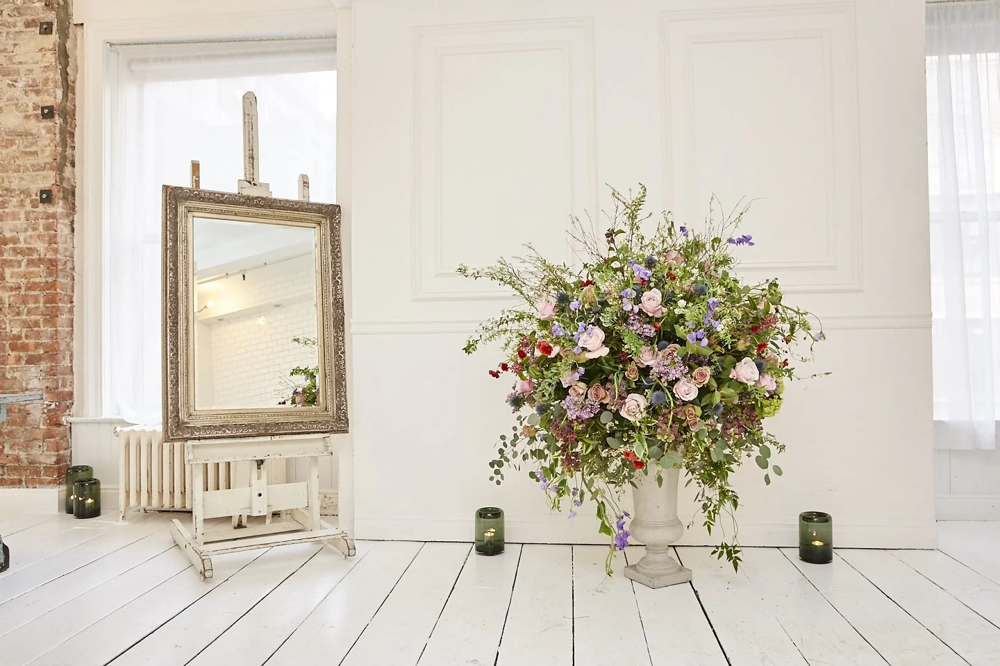 A large floral arrangement with pink roses, purple flowers, and greenery in a white pedestal vase. An ornate framed mirror is on a white stand nearby, with three green candle holders on the floor around the arrangement, set in a bright, elegant room 