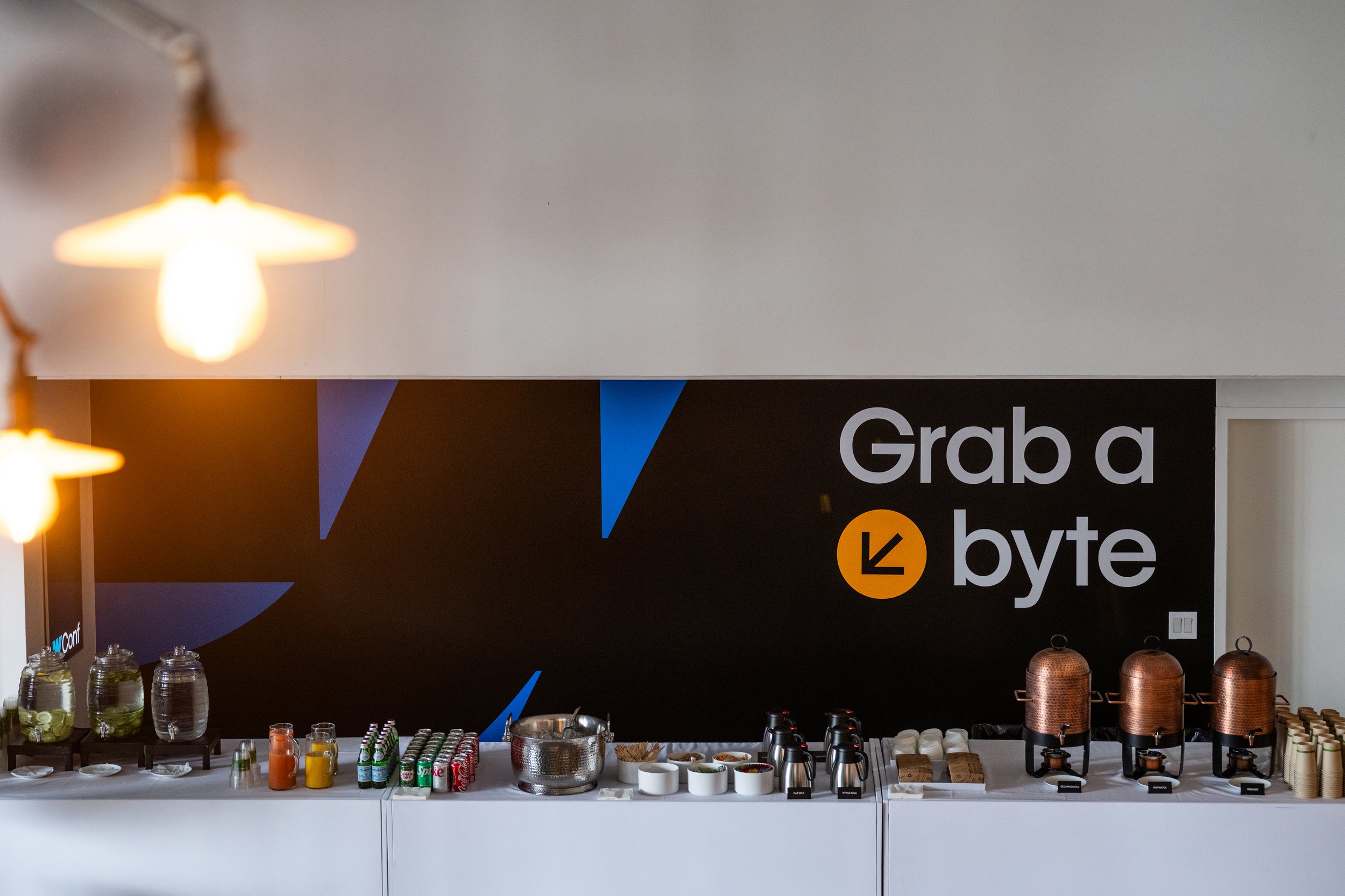 A beverage station with glass jars of infused water, juice bottles, canned drinks, and containers of hot tea in a conference room with a black wall featuring the text 'Grab a byte' and a large arrow icon.
