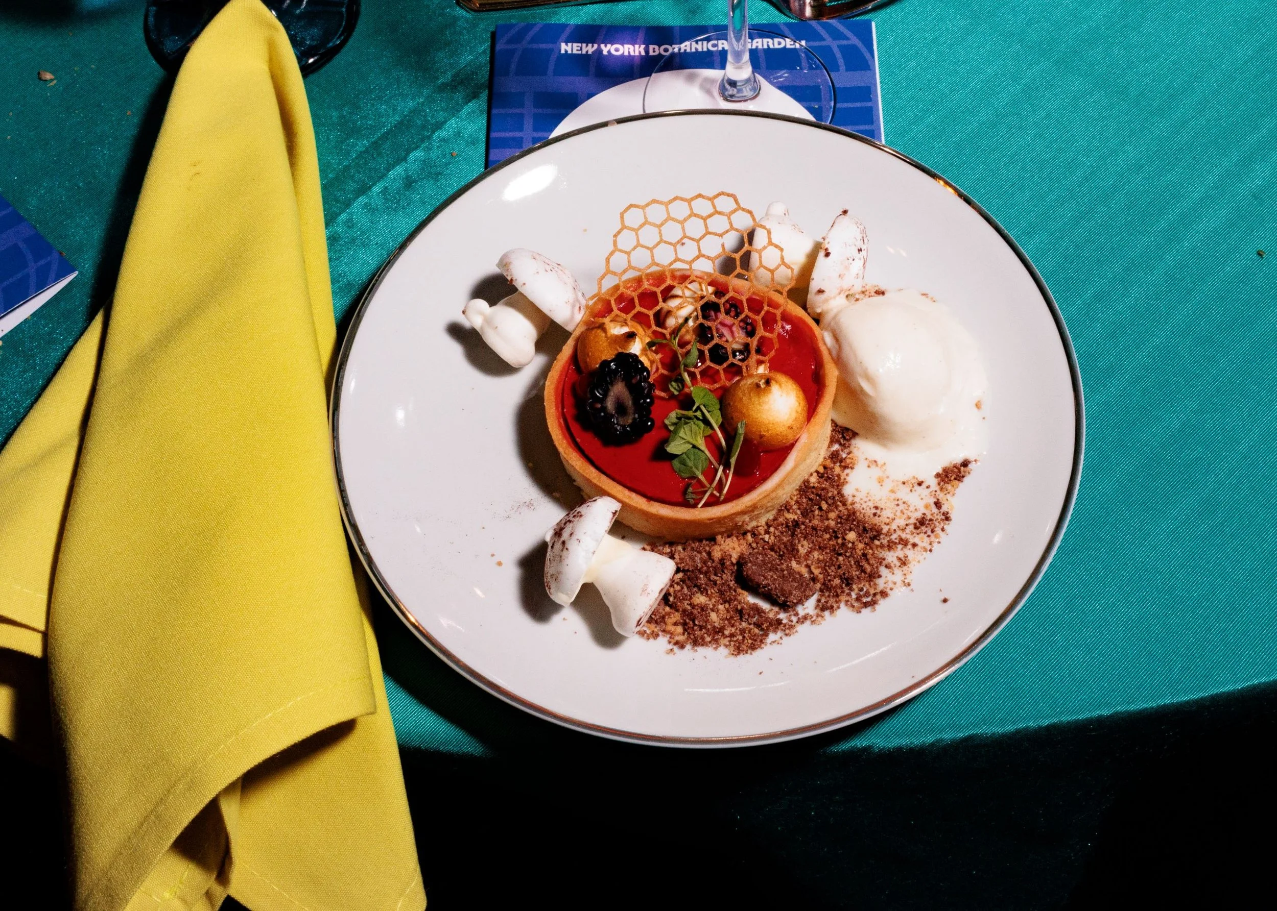 A dessert on a white plate at a table, featuring a tart with berries, a honeycomb piece, a scoop of ice cream, meringue kisses, and powdered cocoa.