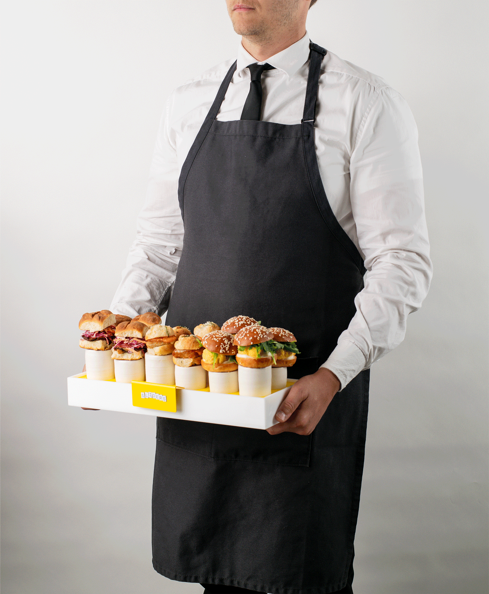 Man in white shirt and black apron holding a tray with mini sandwiches and burgers.