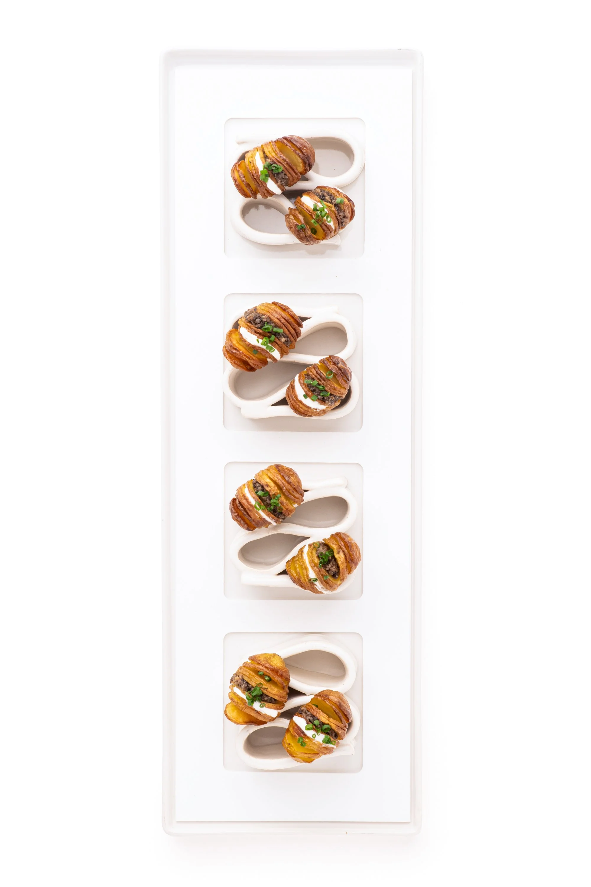 A white rectangular serving tray with four small square compartments, each holding three mini breaded chicken drumsticks garnished with chopped herbs and drizzled with sauce.