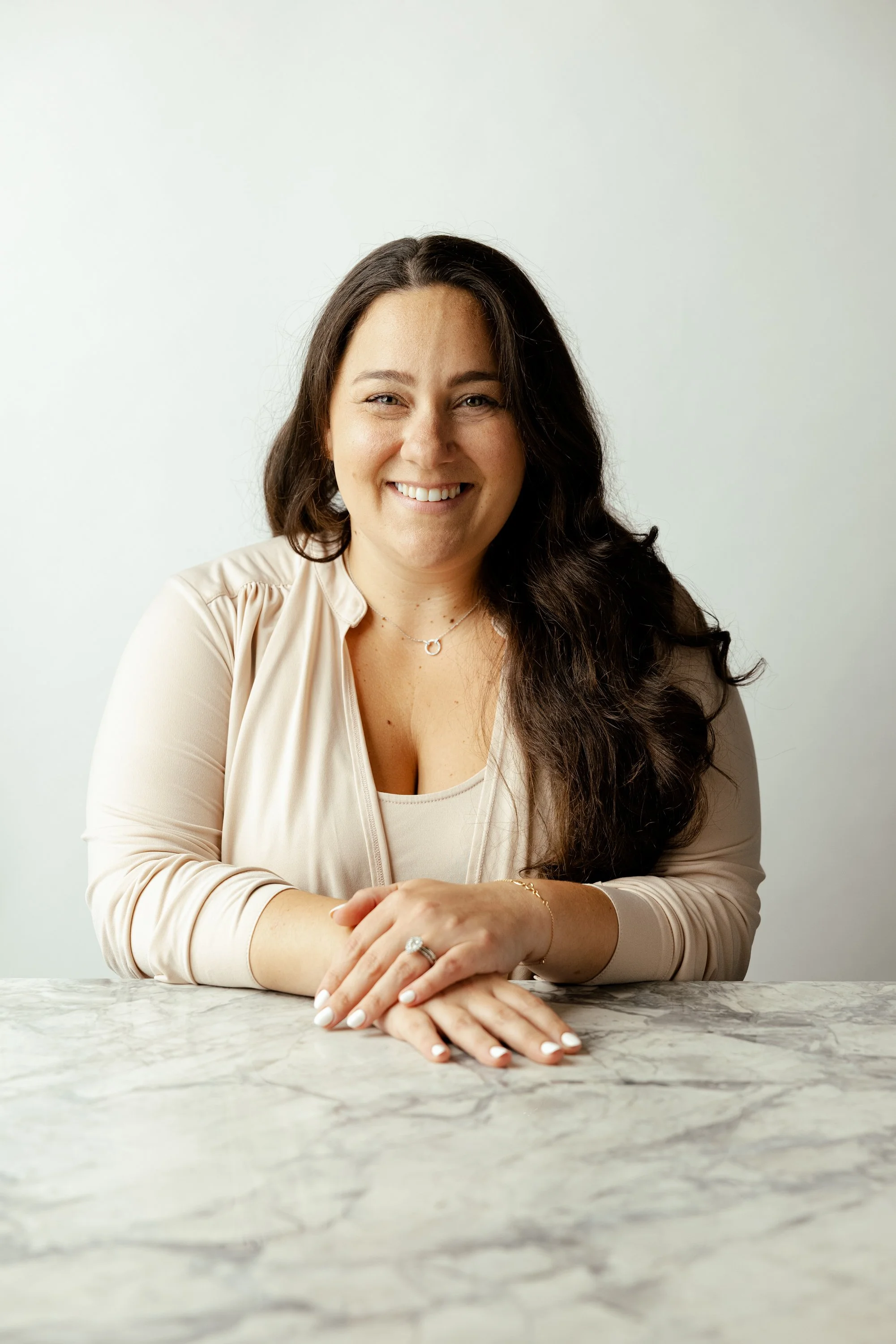 A smiling woman with long dark hair, wearing a beige blazer and jewelry, sitting at a marble table against a plain white wall.