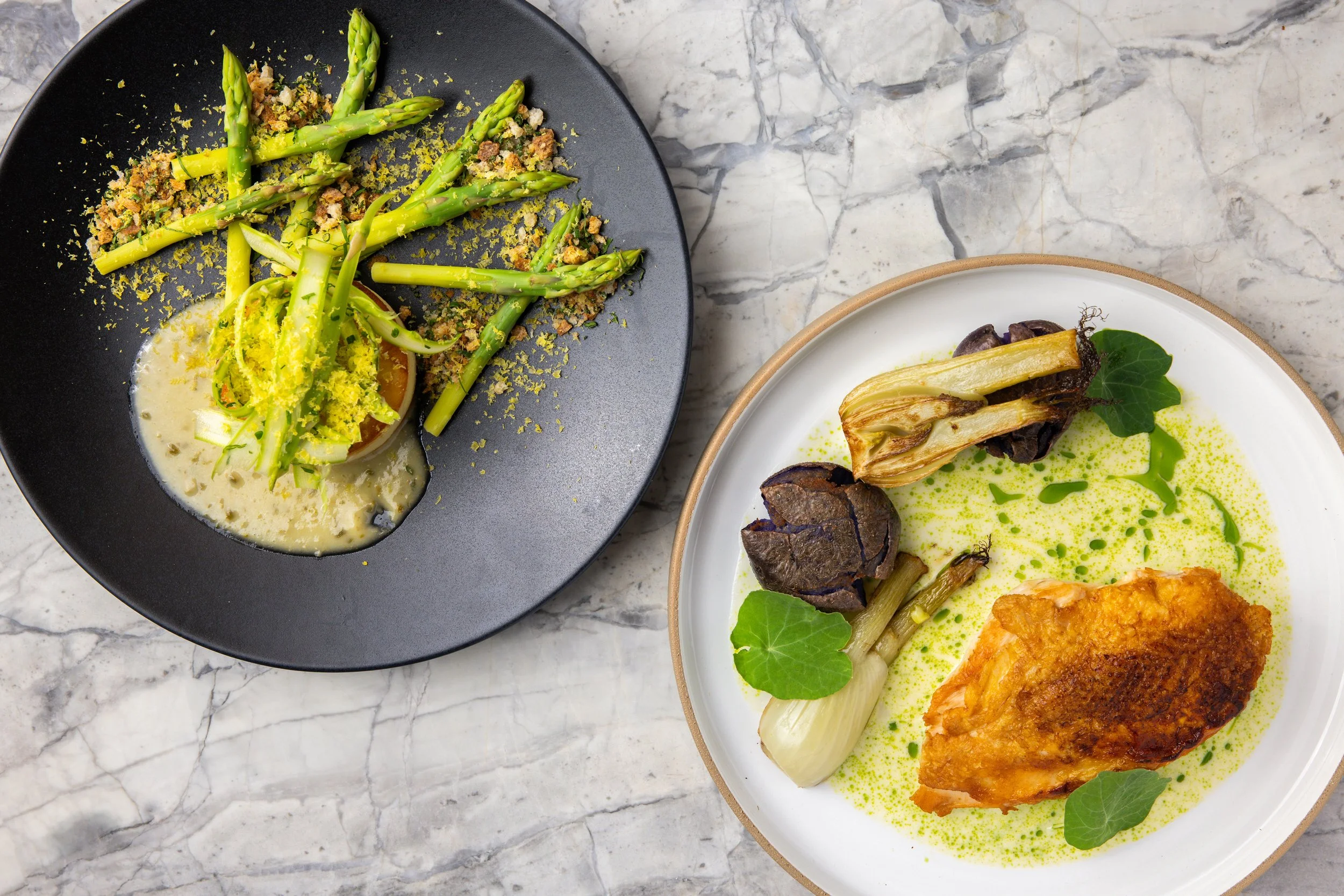 Two plates of gourmet food on a marble surface: the black plate contains white asparagus with a creamy sauce and some green garnish, and the white plate has a fried piece of fish with grilled vegetables and green sauce.