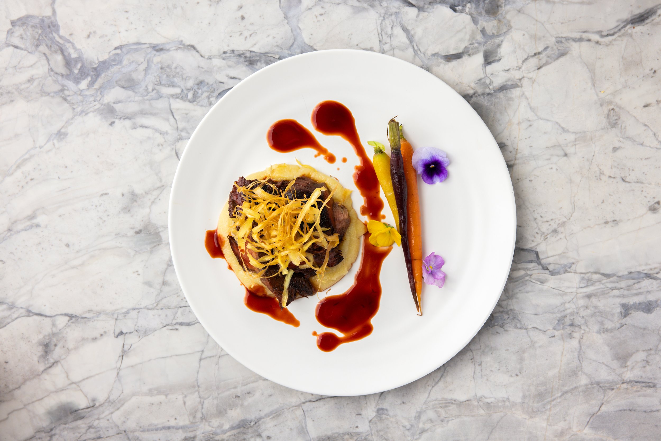 Dish with mashed potatoes topped with shredded cheese and beef, garnished with a dark sauce, on a white plate. Side of baby carrots in different colors and purple edible flowers. The plate is on a light gray marble surface.